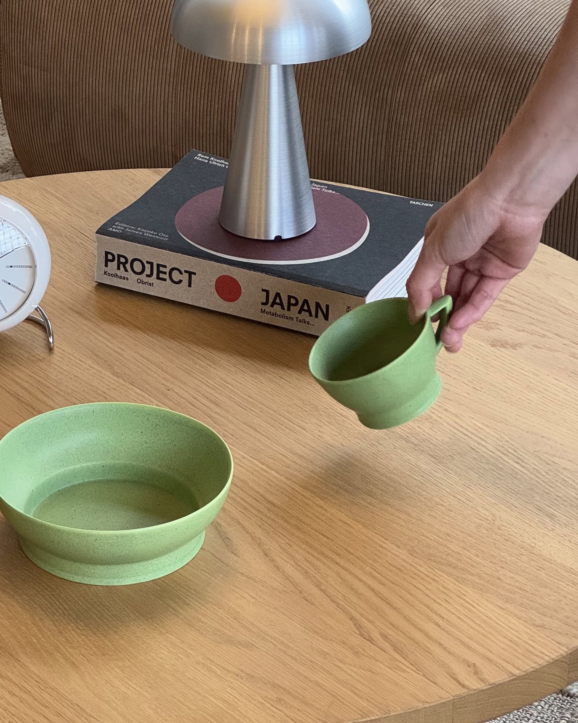 A hand picks up the Serax Ra Cup - Green, a porcelain cup by Serax, from a wooden table that also holds a matching bowl, a clock, a lamp, and the book Project Japan.