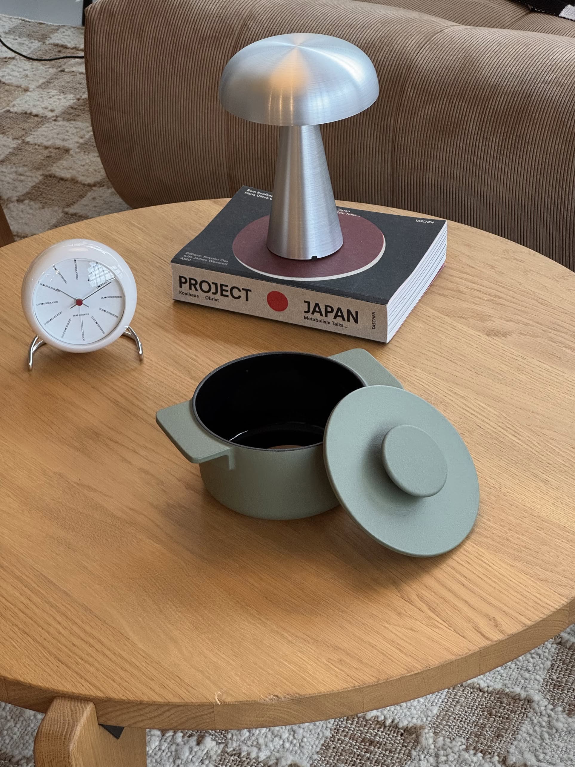 A round wooden table displays the Serax Surface Cast Iron Pot XS - Camogreen, a modern clock, and books topped with a metallic mushroom lamp. A brown striped chair and patterned carpet in the background evoke cozy kitchen vibes.