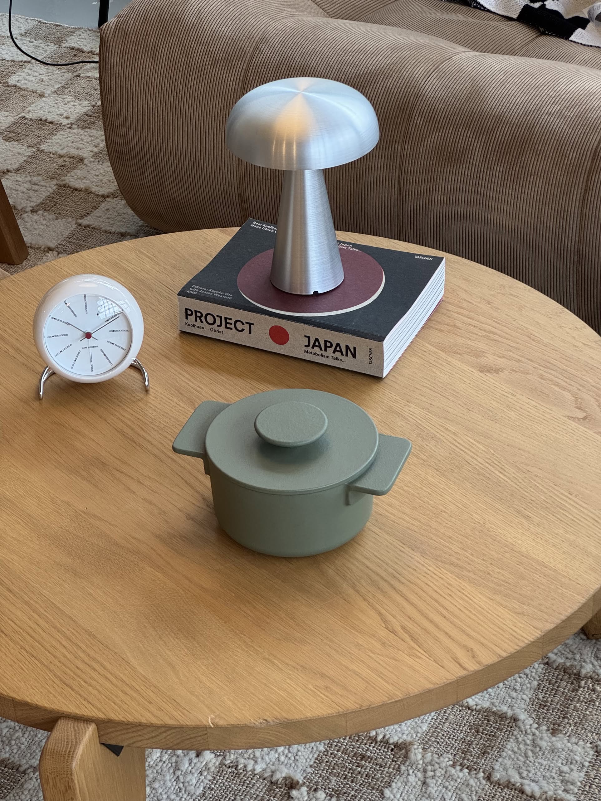 A round wooden table features the Serax Surface Cast Iron Pot XS - Camogreen, a silver mushroom-shaped lamp atop two books, and a white clock. In the background, a brown sofa and patterned rug add to the cozy, kitchen-inspired atmosphere.