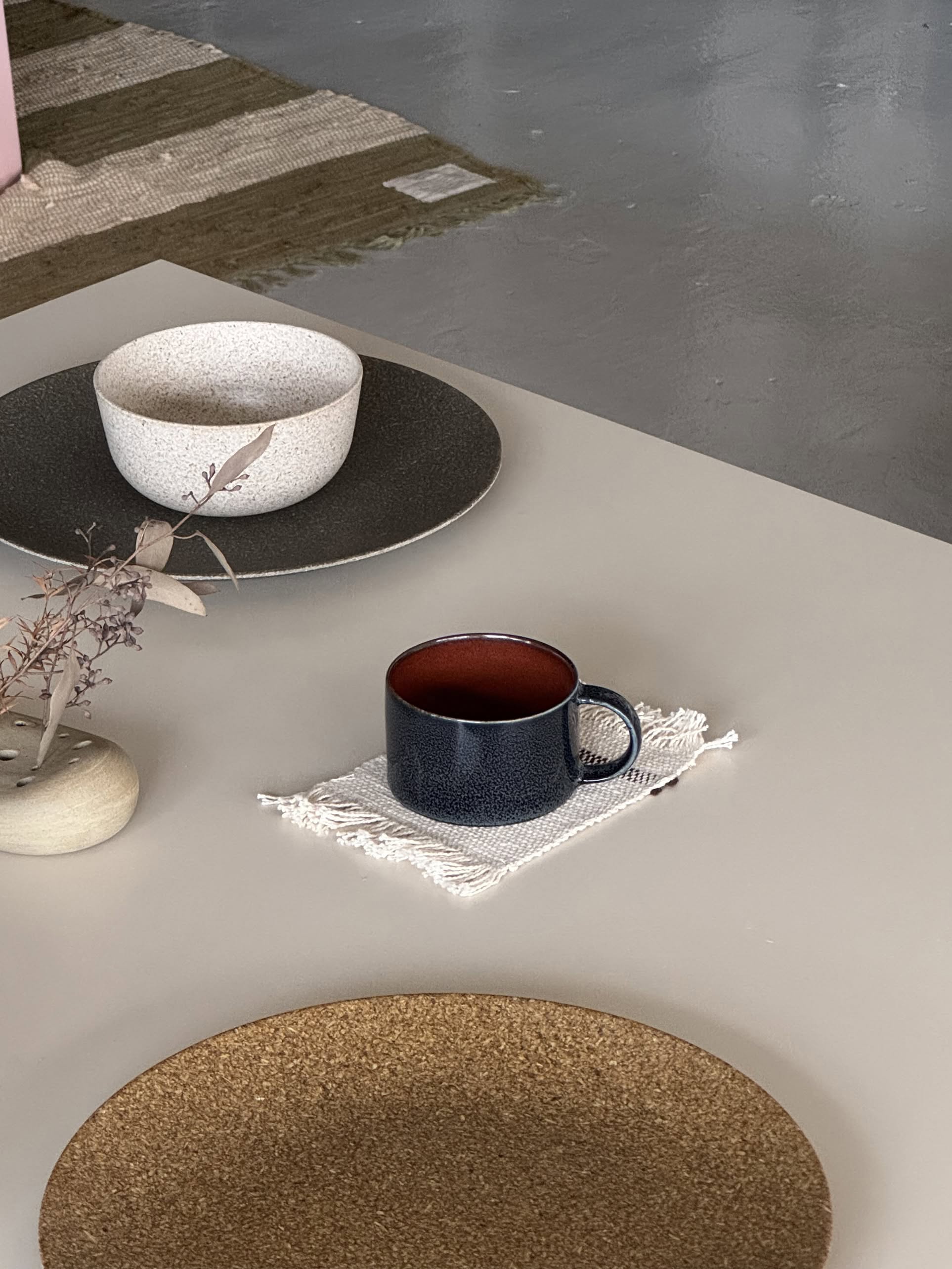 The Serax Terres De Rêves Coffee Cup by Serax sits on a white fringed cloth atop a light table, paired with a beige bowl, brown placemat, and small vase of dried leaves. A textured rug covers the gray floor in the background.