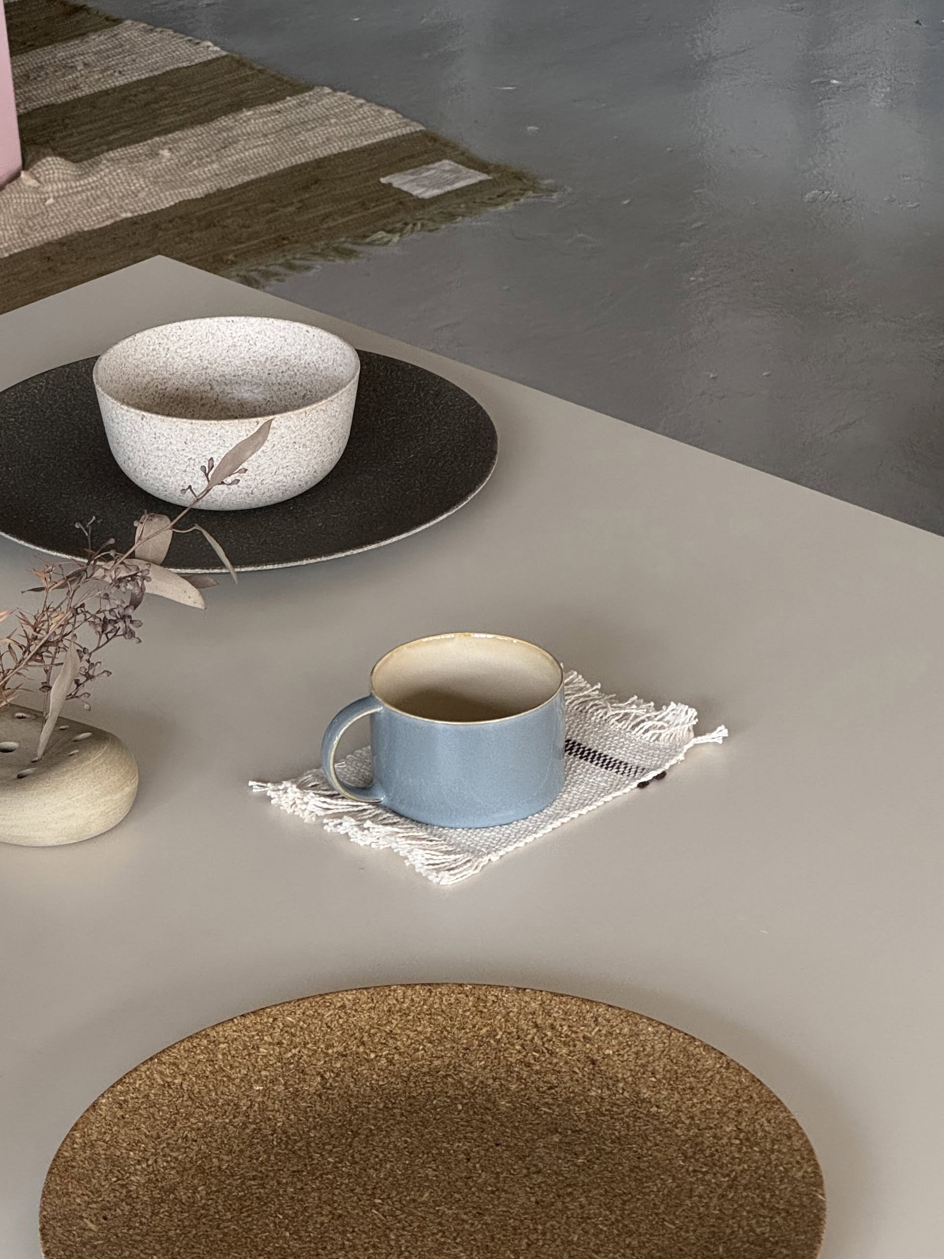 A minimalist table setting features the Serax Terres De Rêves Coffee Cup by Serax on a white cloth coaster, a gray bowl on a dark plate, a beige bowl on a cork mat, and a simple vase with dried branches. A rug lies on the polished concrete floor in the background.