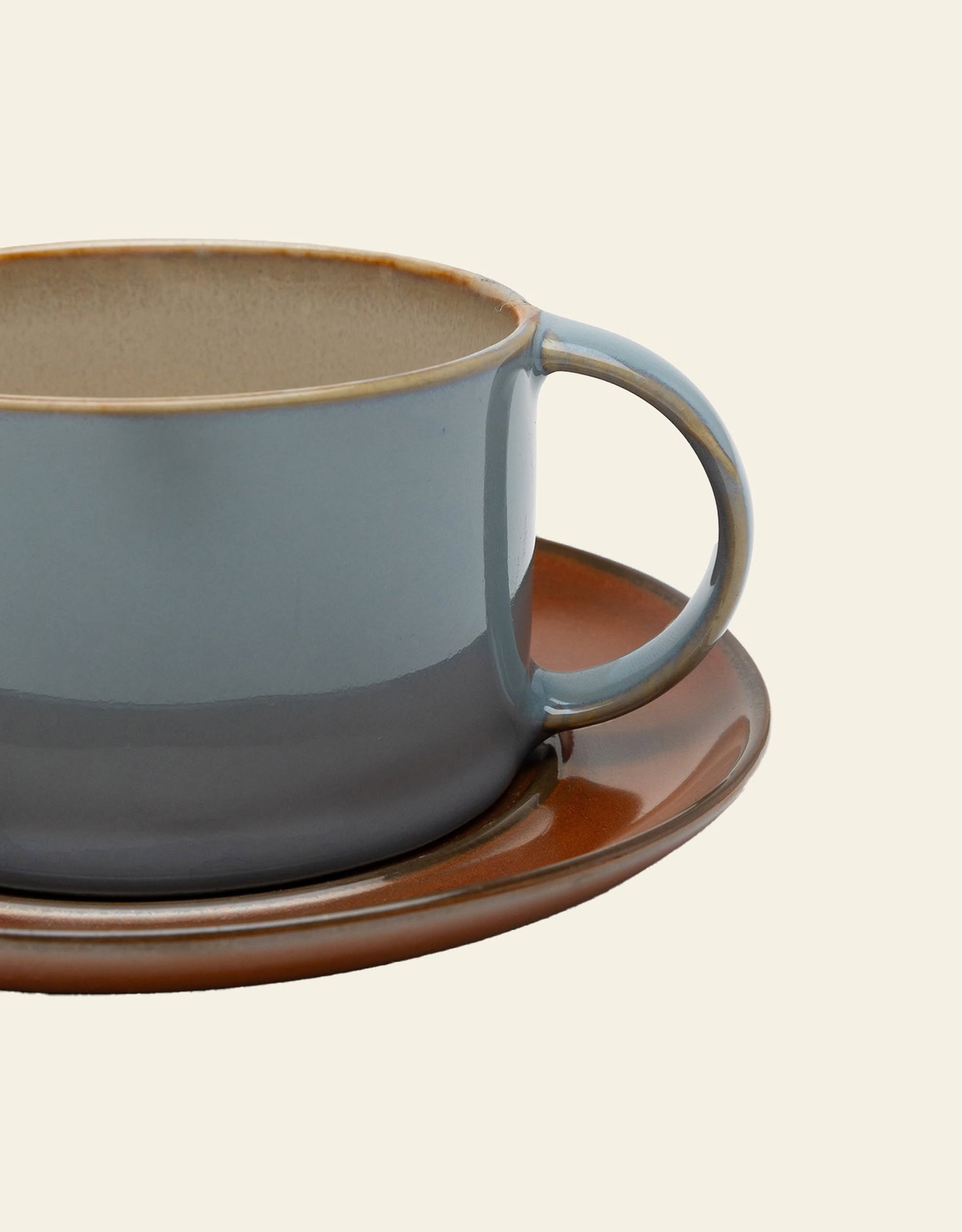 A close-up of the Serax Terres De Rêves Coffee Cup by Serax, showcasing its blue-gray gradient and handle, sits partly visible on a matching brown saucer against a plain beige background.