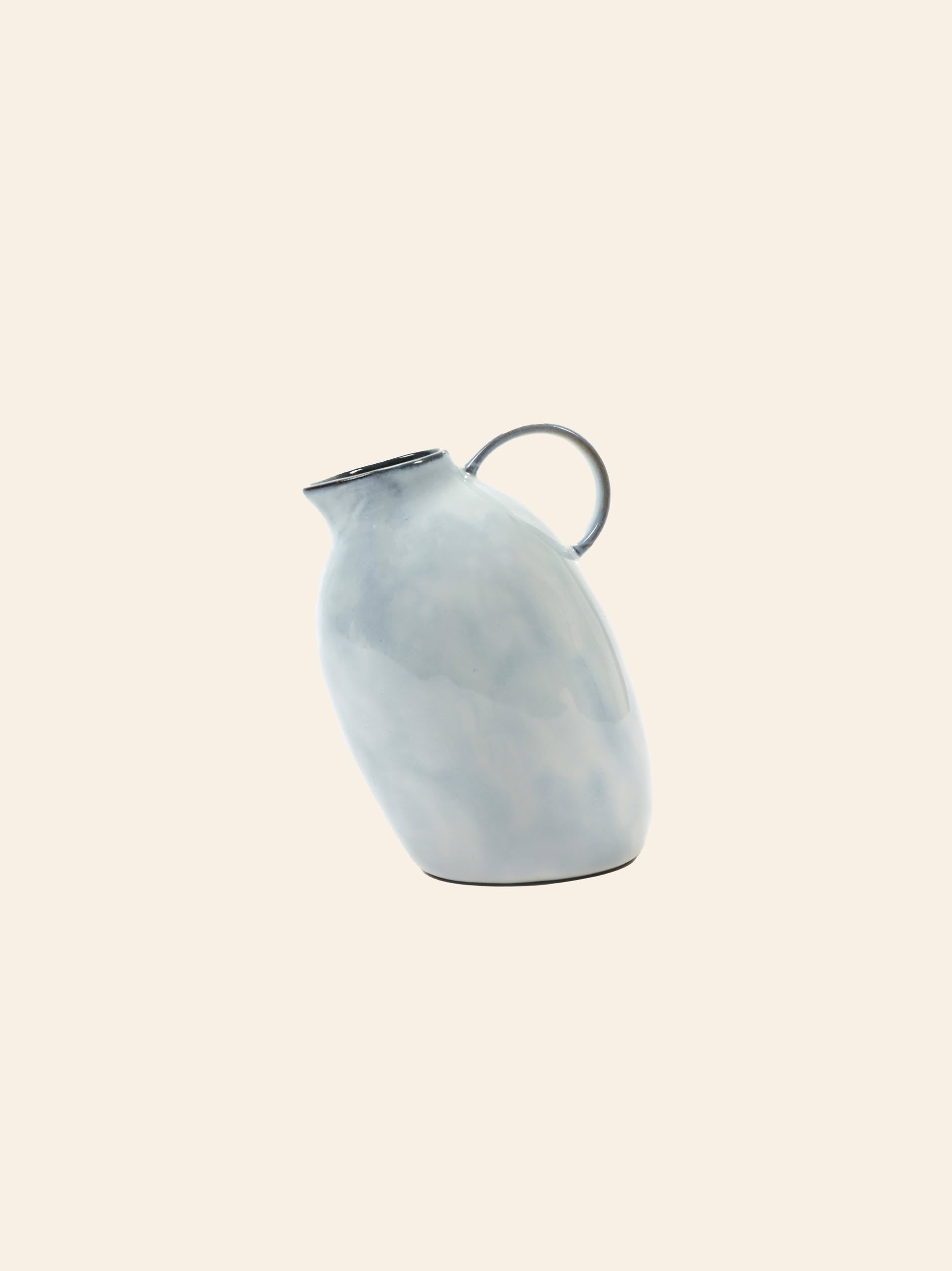 The Serax Terres De Rêves Jug °1 in glossy white ceramic, featuring a round handle and oval-tilted body, stands out as a unique design piece by Serax, showcased against a plain beige backdrop.
