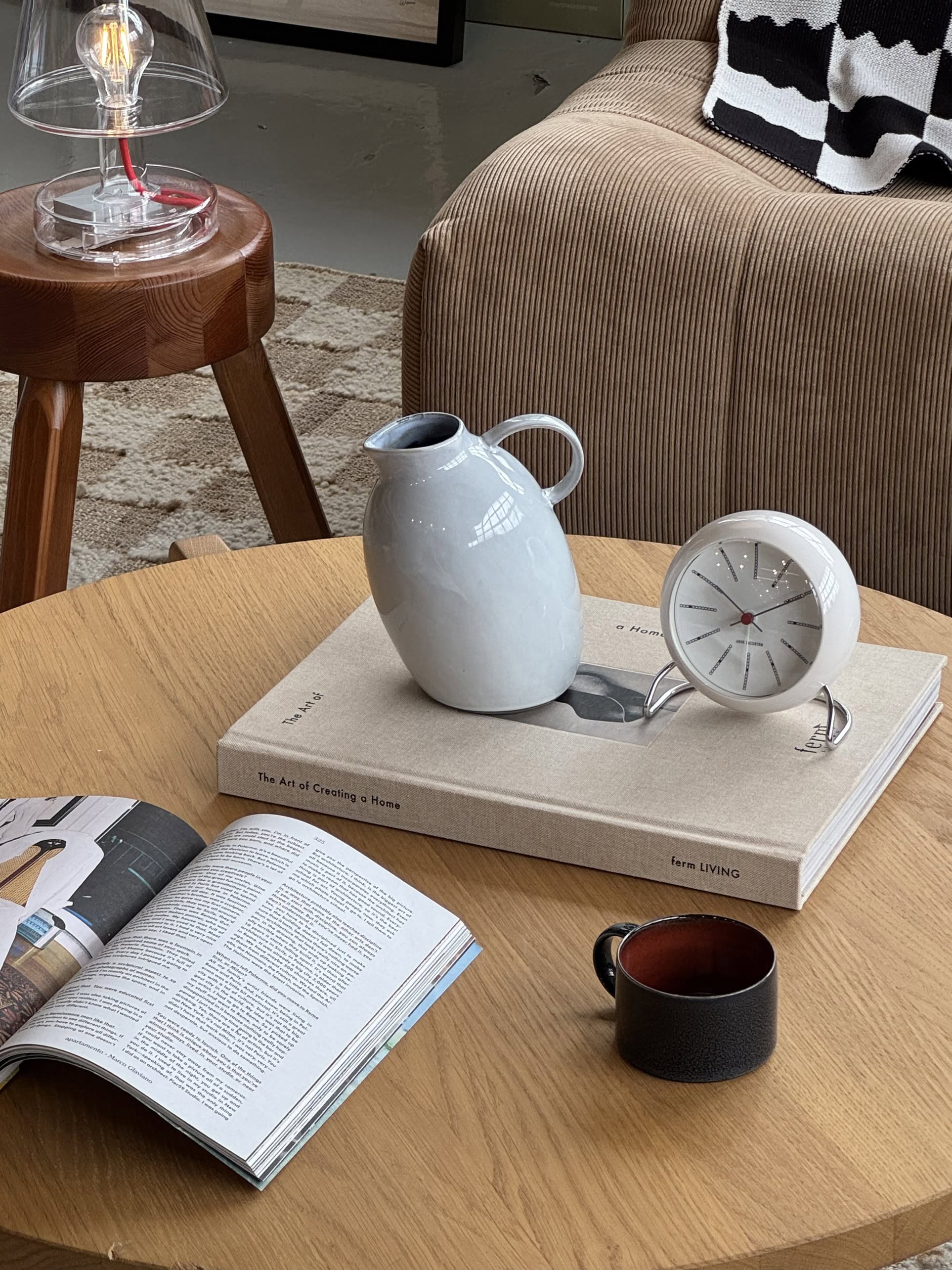 A wooden coffee table serves as a statement piece, featuring a black cup, open magazine, large book, Serax Terres De Rêves Jug °1 - White by Serax, white clock, and a beige armchair with a black and white blanket in the background.