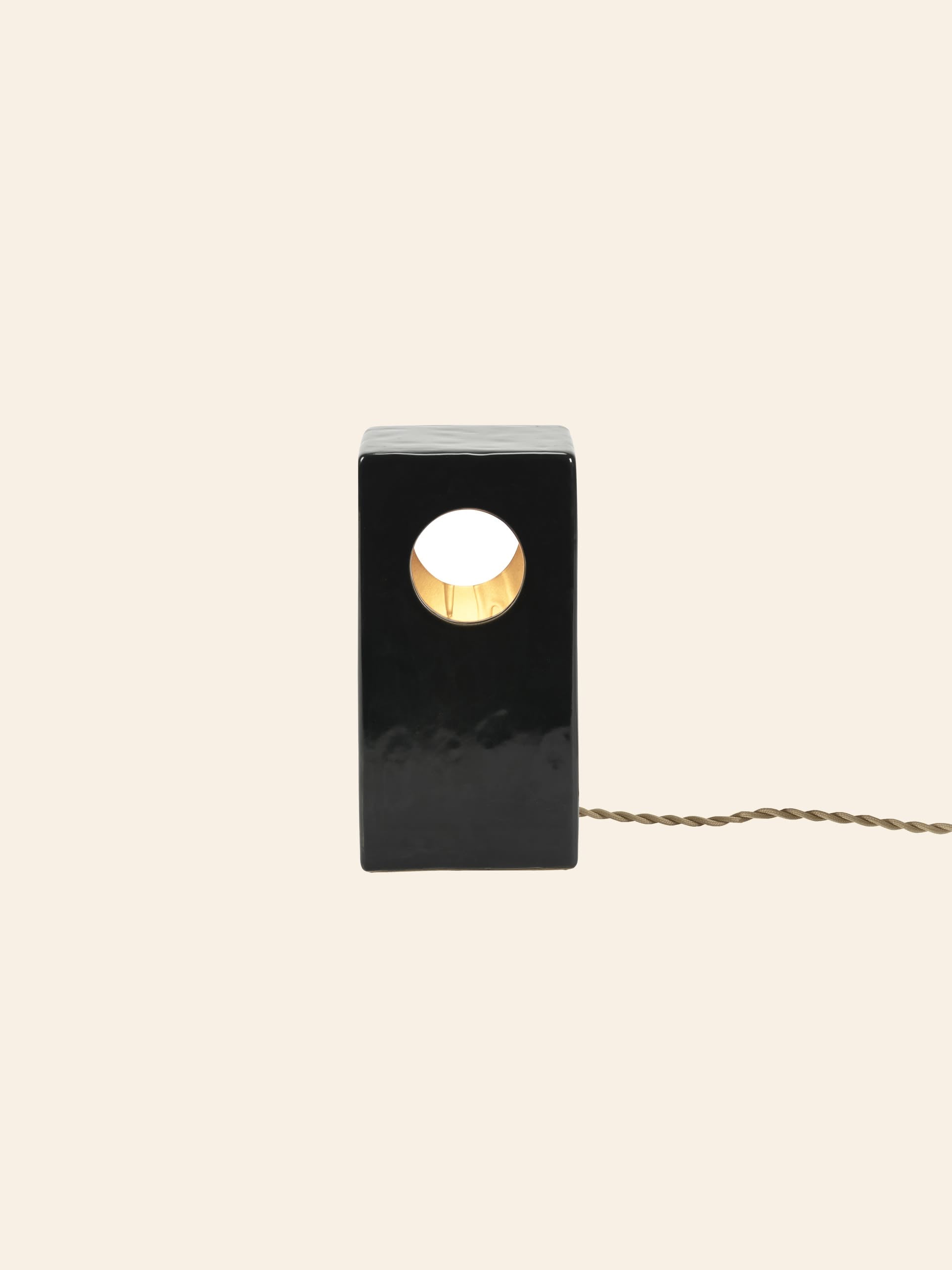 The Serax Terres De Rêves Maxime N°1 Table Lamp in black features a rectangular stoneware base with a central circular hole, emitting warm light. Its geometric design and twisted cord stand out against a plain beige background.