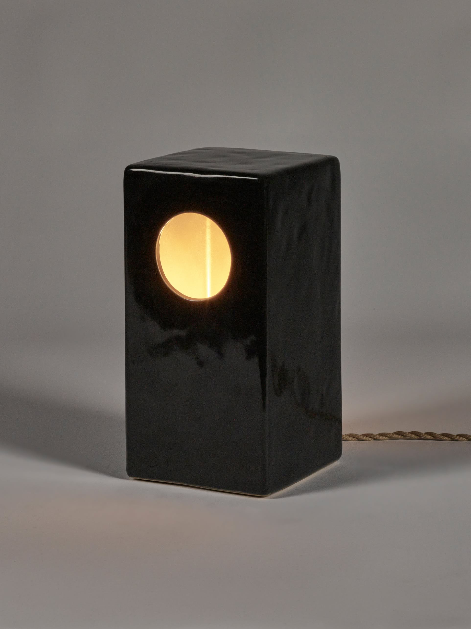 The Serax Terres De Rêves Maxime N°1 Table Lamp in black stoneware features a glossy rectangular body with a circular geometric design, emits warm yellow light, and has a twisted power cord extending from the back.