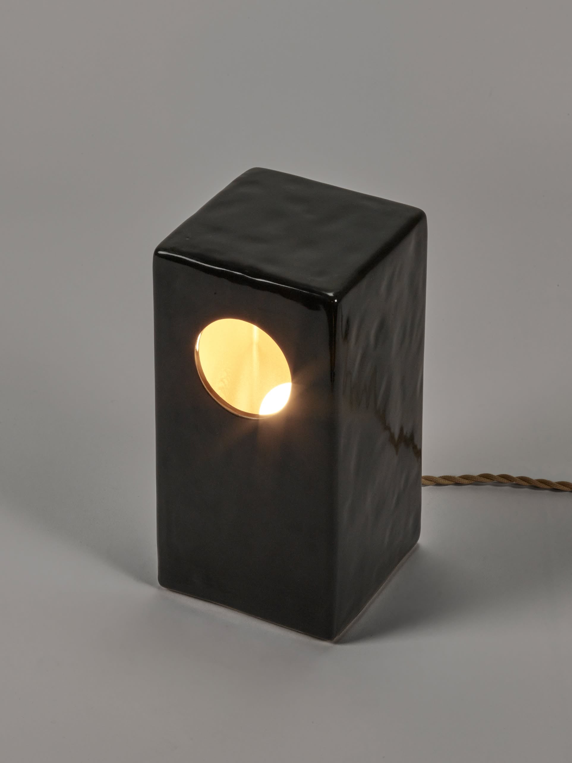 The Serax Terres De Rêves Maxime N°1 Table Lamp in black features a rectangular stoneware body with geometric design, a circular cutout for warm light, textured surface, and a visible power cord for modern style.