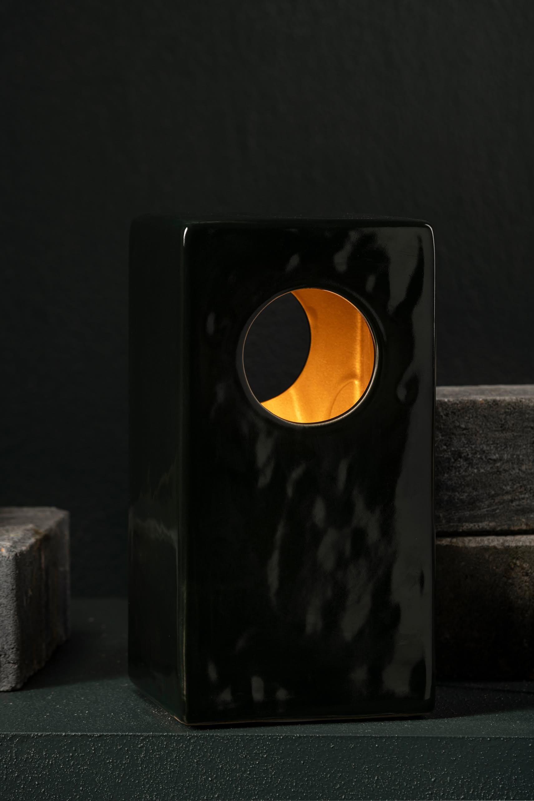 The Serax Terres De Rêves Maxime N°1 Table Lamp in glossy black stoneware features a circular cutout and geometric design, emitting a warm yellow-orange glow against a dark background, with dark stone blocks on either side.