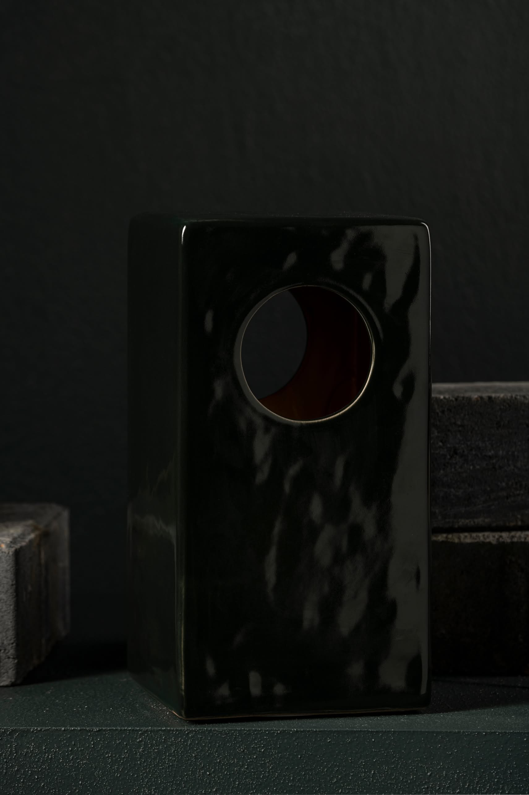 The Serax Terres De Rêves Maxime N°1 Table Lamp in glossy black stoneware features a rectangular shape with a round top hole, standing out against dark bricks and a black wall.