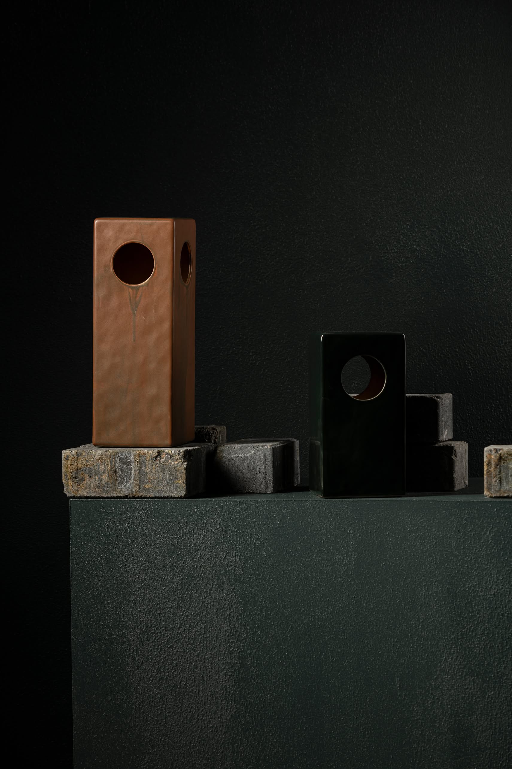 The Serax Terres De Rêves Maxime N°1 Table Lamp by Serax features a geometric stoneware design in black, showcased on stacked stone blocks against a dark textured background with dramatic lighting that highlights its bold form.