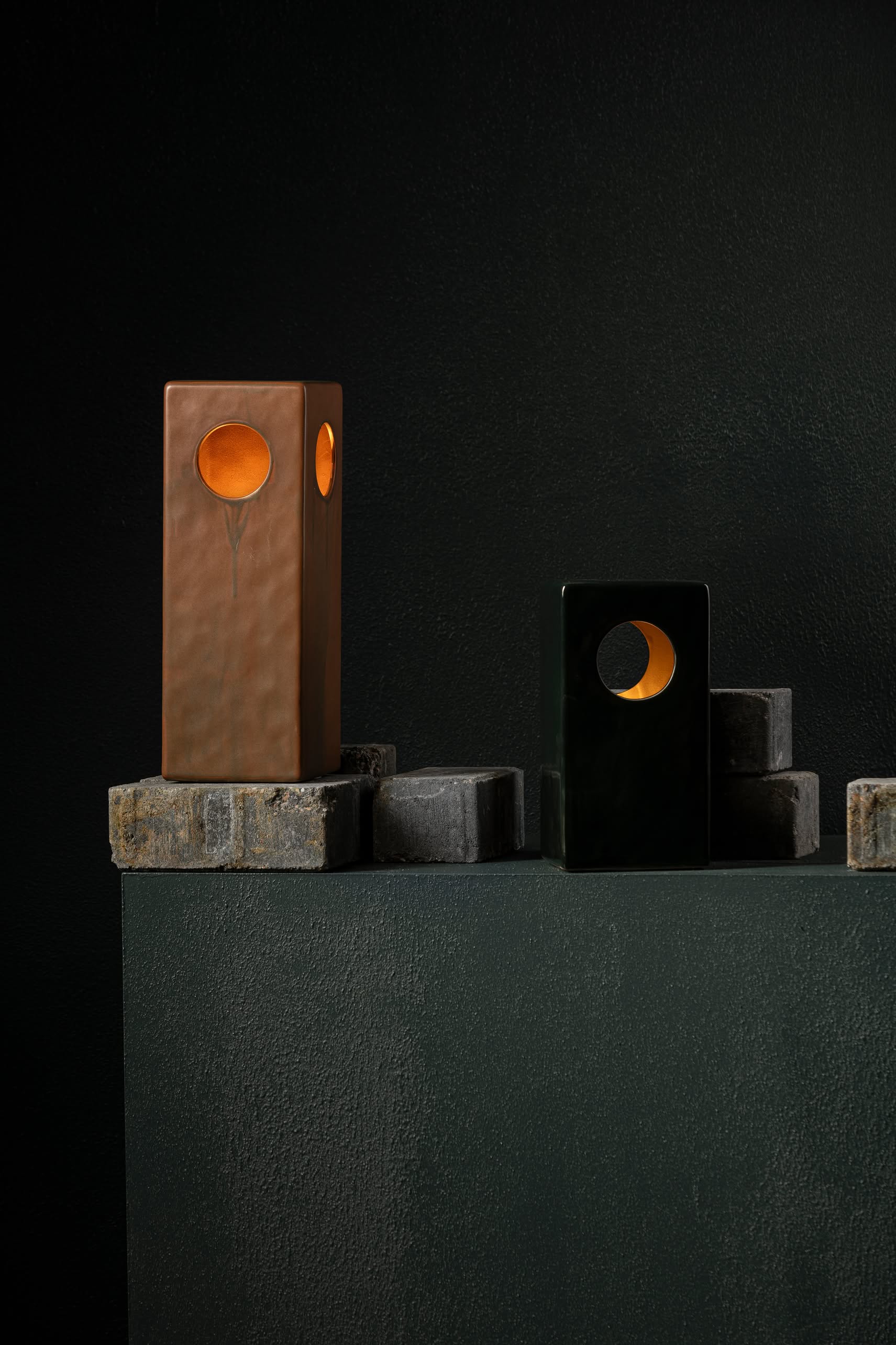 The Serax Terres De Rêves Maxime N°1 Table Lamp in black stands on stacked stoneware blocks, its geometric design and circular cutout glowing warmly against a dark, textured background.