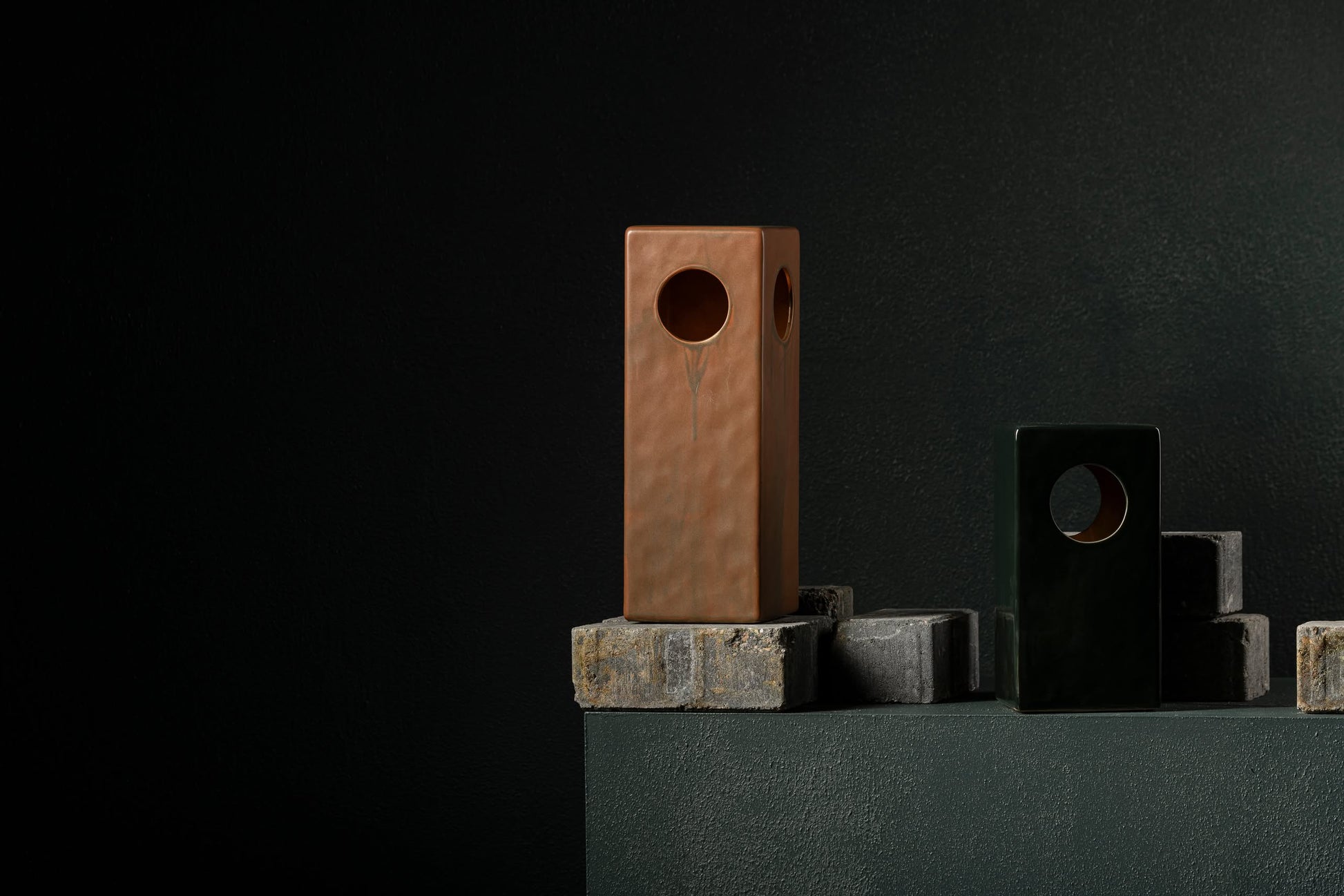 The Serax Terres De Rêves Maxime N°1 Table Lamp in black by Serax features a striking geometric design, displayed on gray concrete blocks against a dark textured background to highlight its unique circular cutout.