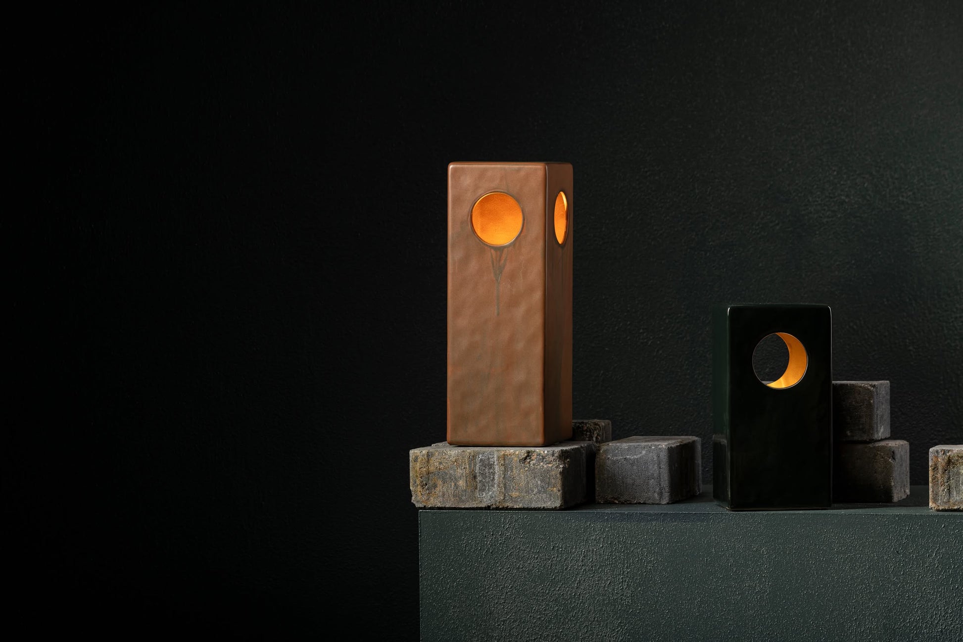Two Serax Terres De Rêves Maxime N°1 Black stoneware table lamps with circular cutouts emit a warm orange glow. Their geometric design on gray stone blocks against a dark textured wall creates a modern, minimalist setting.