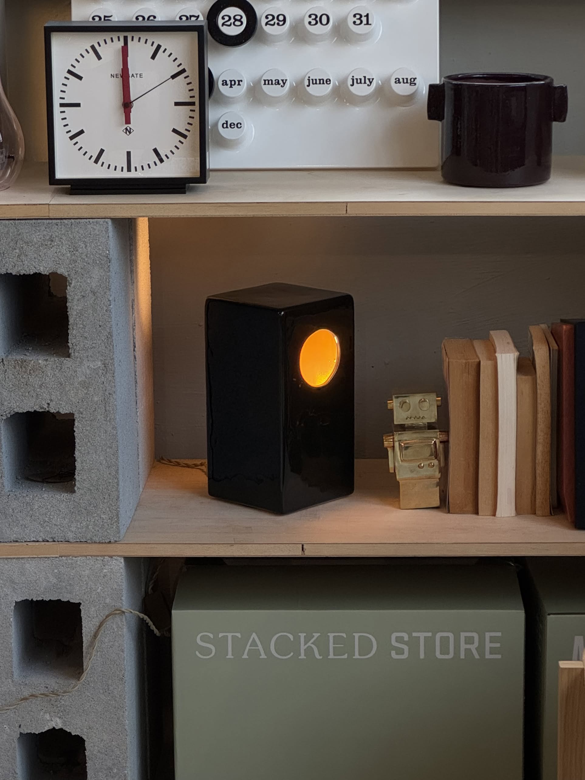 A small shelf displays a black clock, a white calendar, the Serax Terres De Rêves Maxime N°1 Table Lamp in black with an orange glow, a gold robot figurine, books, and a STACKED STORE box, divided by gray cinder blocks.
