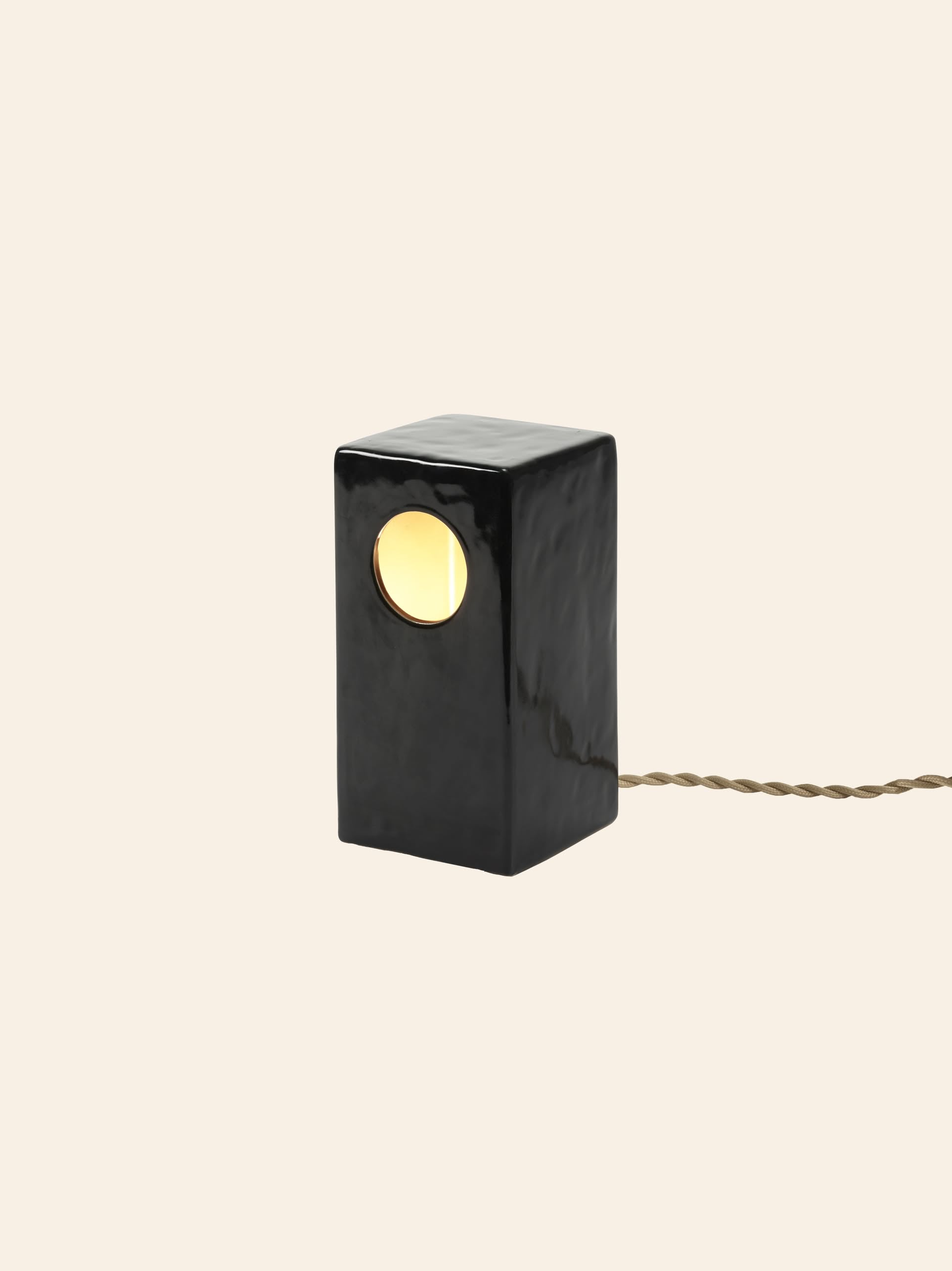 The Serax Terres De Rêves Maxime N°1 Table Lamp in black features a minimalist stoneware design with a rectangular geometric shape, a round illuminated opening, and a twisted beige power cord. Brand: Serax.
