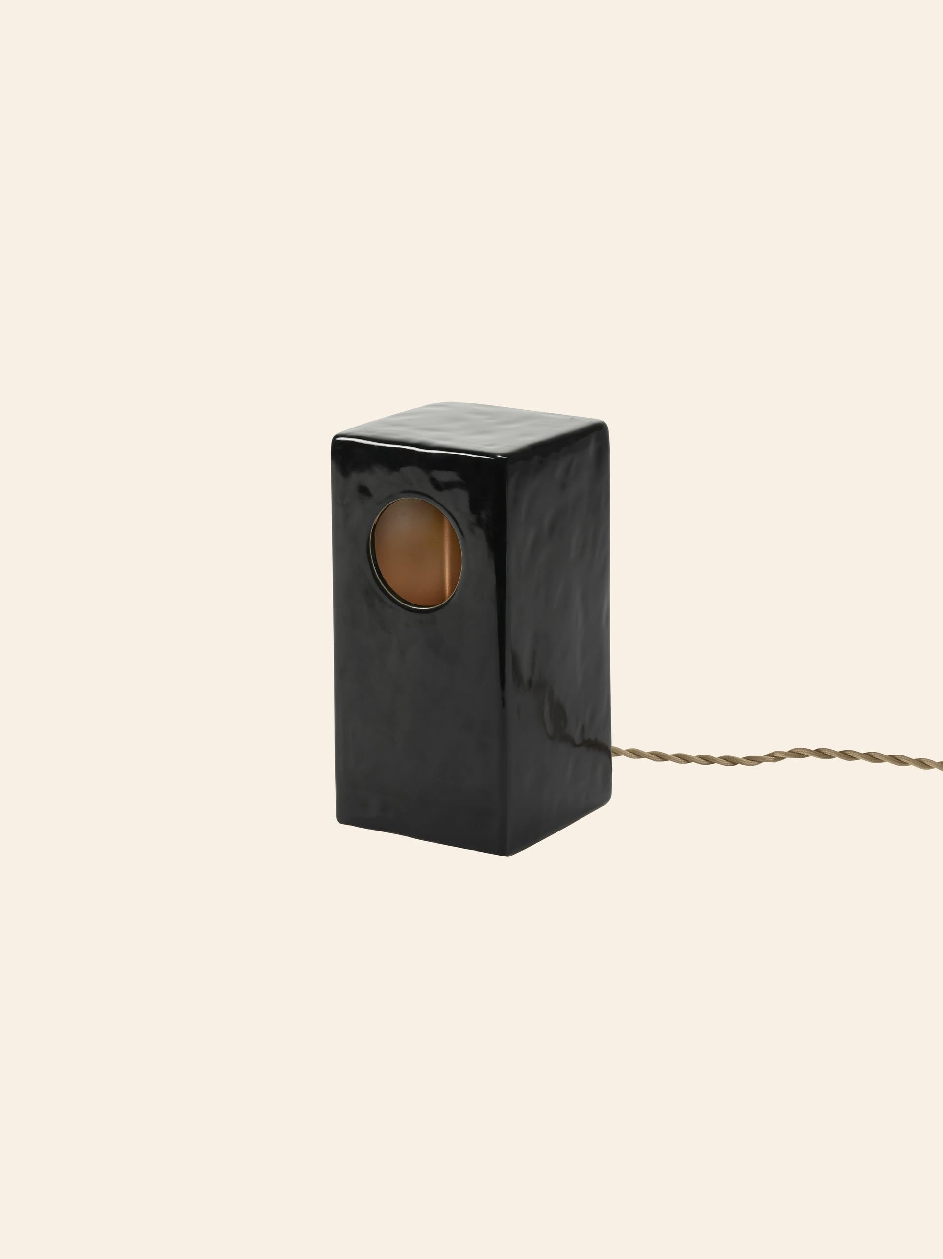 The Serax Terres De Rêves Maxime N°1 Table Lamp is a glossy black, geometric stoneware lamp with a circular opening for warm light and a twisted brown cord, shown against a beige background.