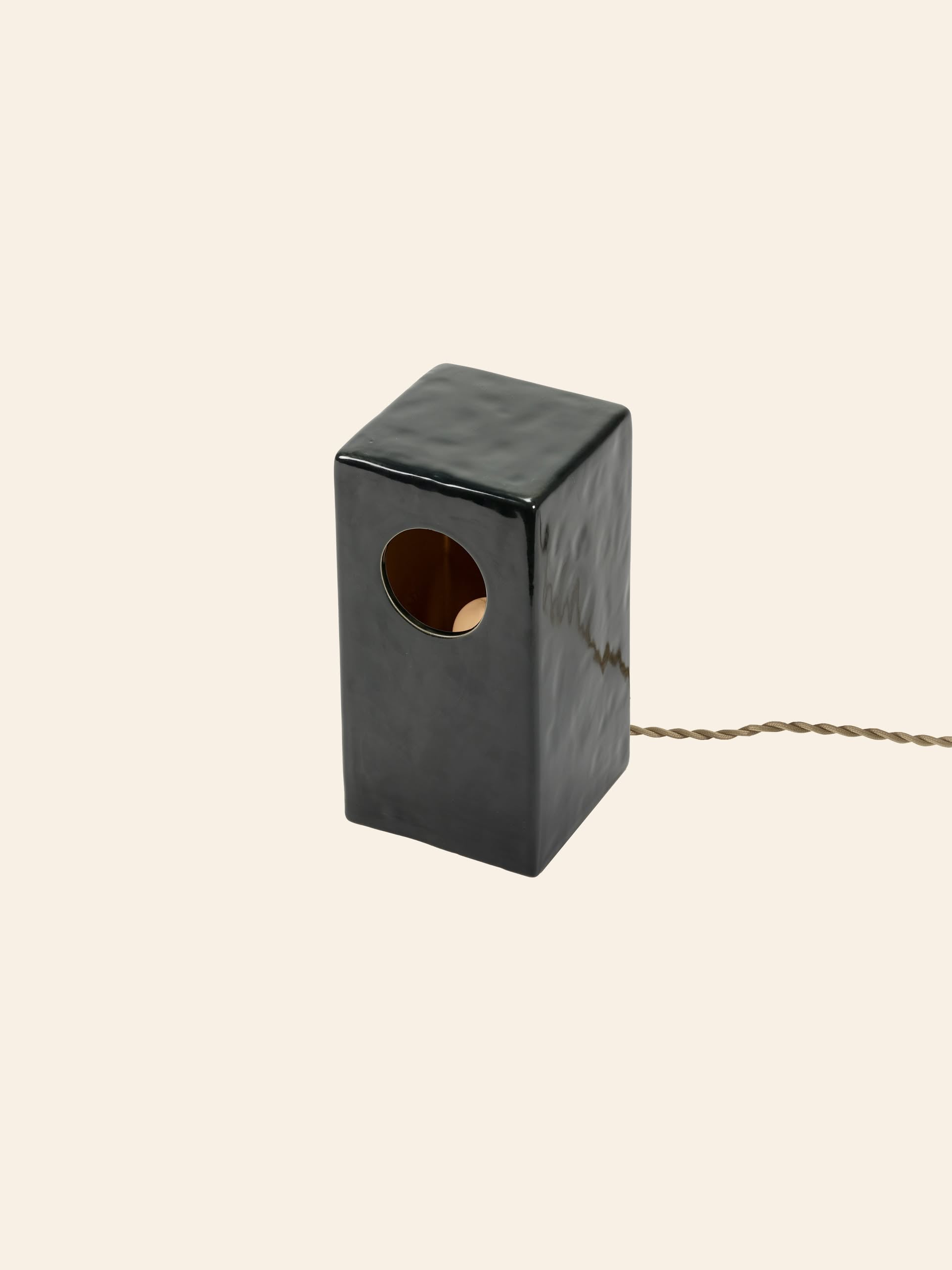 The Serax Terres De Rêves Maxime N°1 Table Lamp in glossy black stoneware features a geometric rectangular design with a round side hole and twisted brown cord, by Serax, shown against a plain beige background.