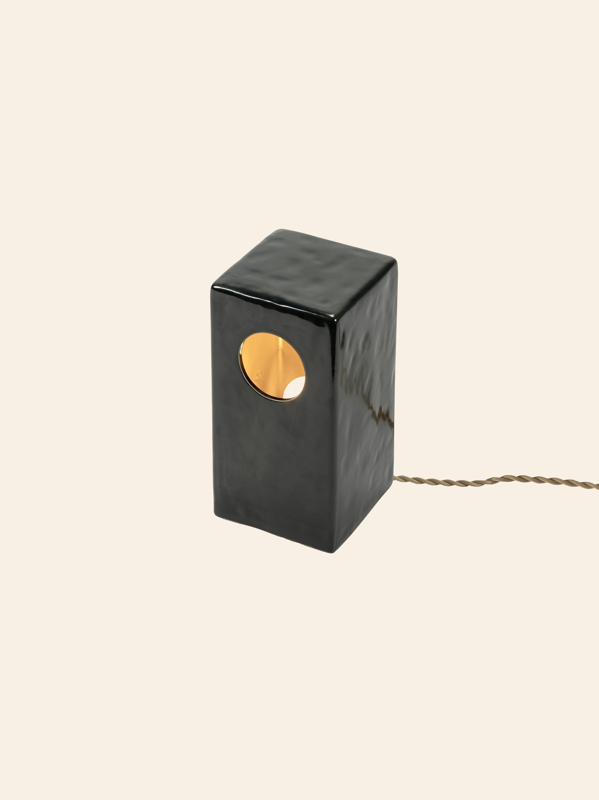 The Serax Terres De Rêves Maxime N°1 Table Lamp by Serax features a modern rectangular black stoneware shape with a circular cutout and twisted brown cord, softly glowing against a plain beige backdrop.