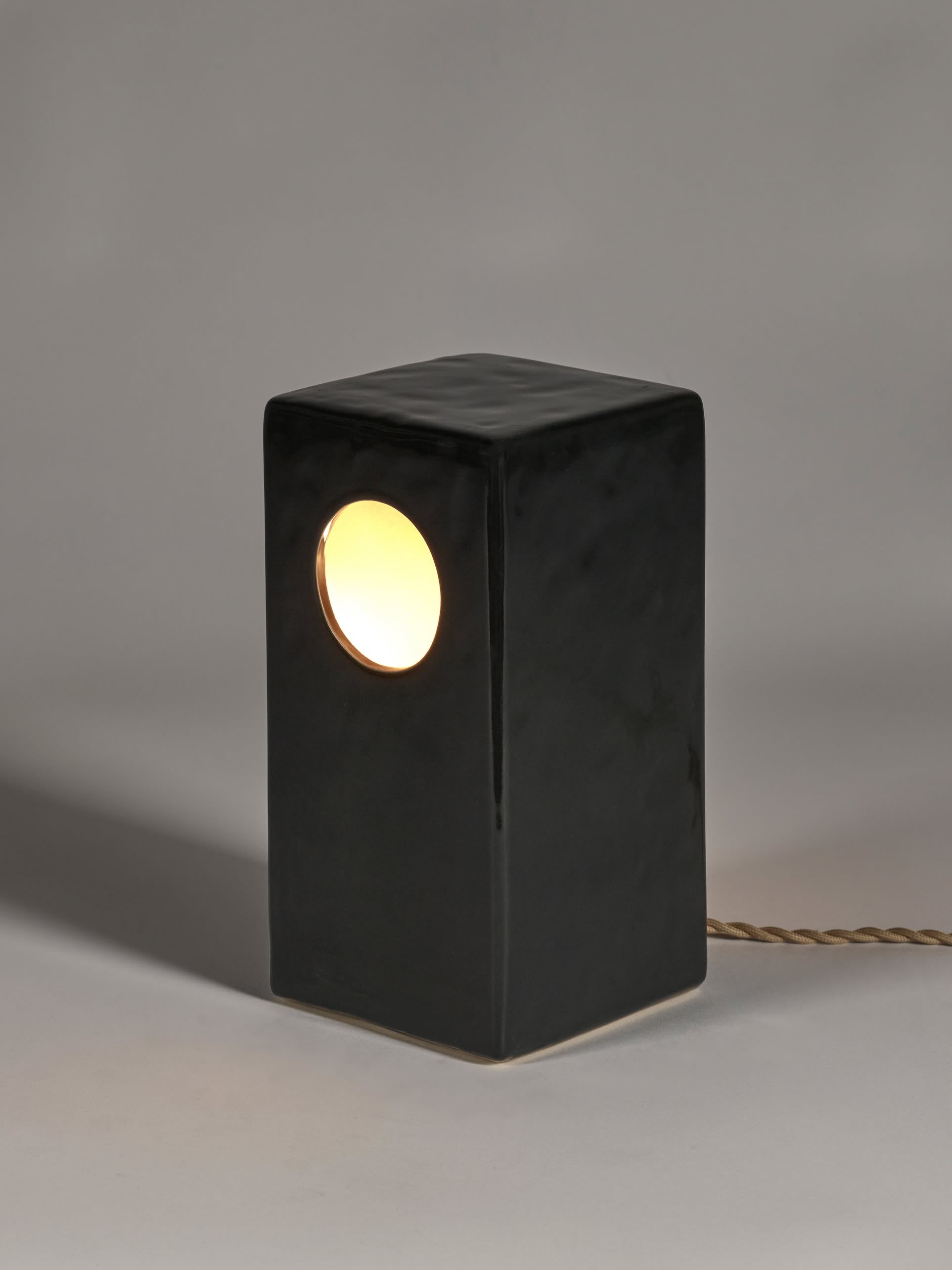 The Serax Terres De Rêves Maxime N°1 Table Lamp in black features a rectangular stoneware base with a round side hole, warm light, and a twisted fabric cord. Its geometric Serax design stands out on a neutral gray surface with a soft shadow.