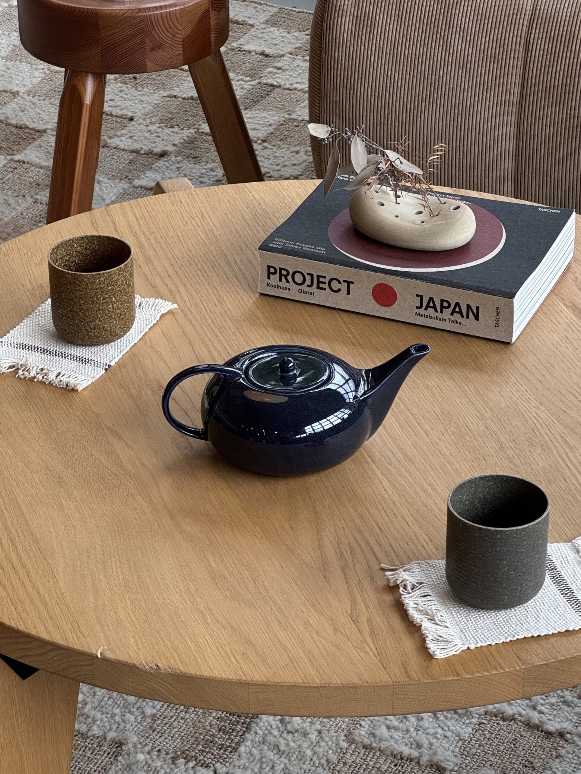 A round wooden table features a Serax Terres De Rêves Teapot - Dark Blue at the center, three elegant pieces of tableware on fabric coasters, and two stacked books (one titled PROJECT JAPAN) beside a decorative animal-shaped object.