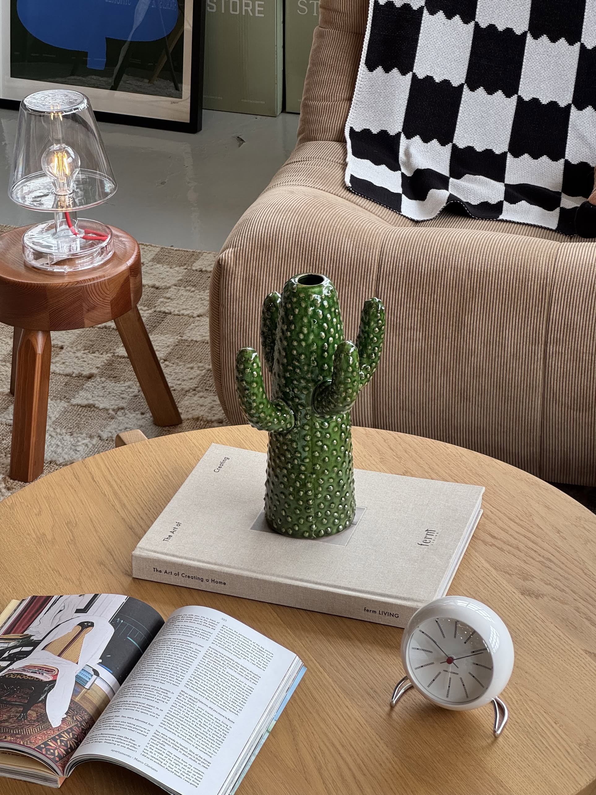 The Serax Urban Jungle Cactus Vase M - Green by Serax sits on a closed book atop a wooden coffee table, accompanied by an open magazine, a white alarm clock, a brown chair with a patterned blanket, and a glass lamp on a wooden stool.