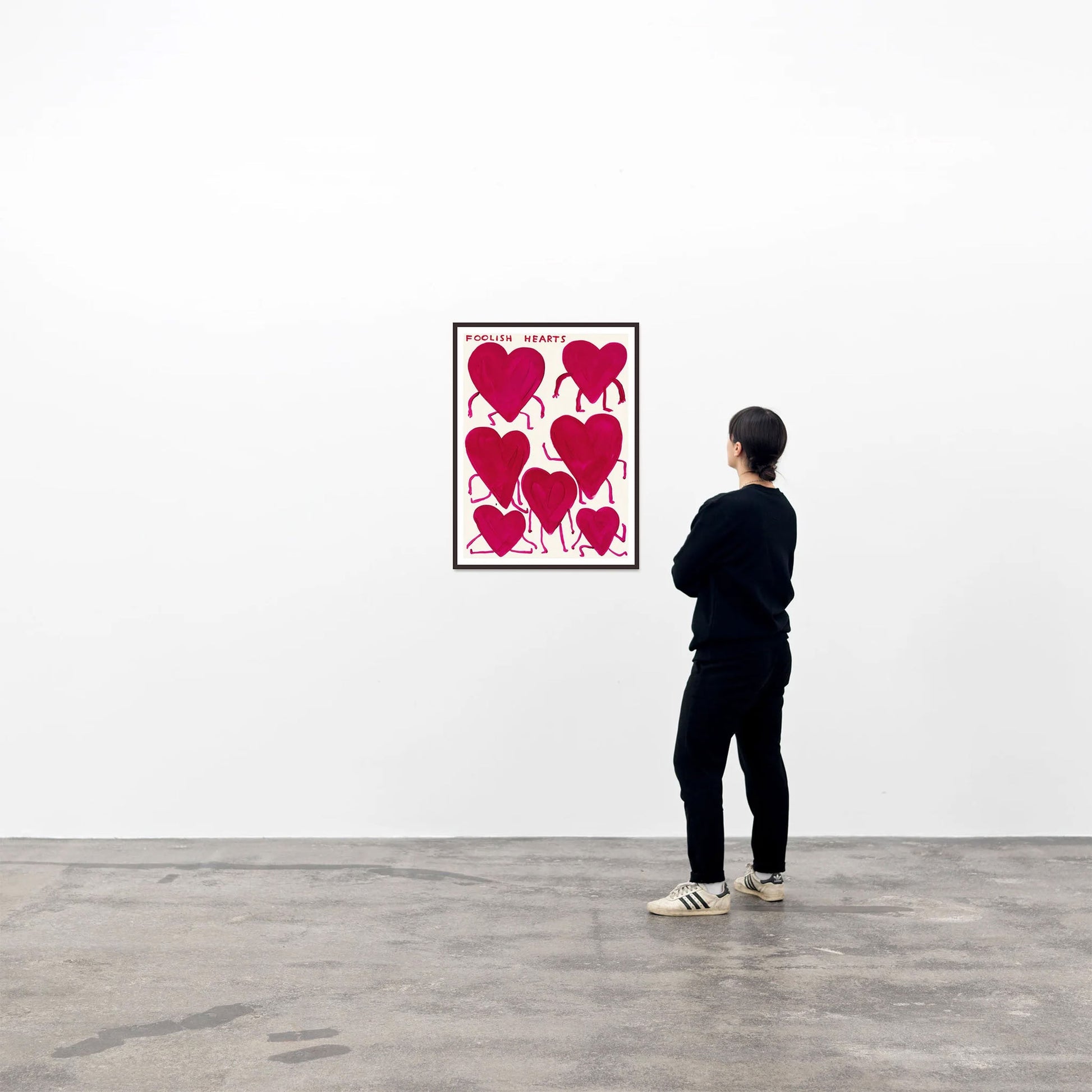 A person in black stands in a gallery, viewing the Shrig Shop David Shrigley - Foolish Hearts (50x70) poster with six red hearts on a white wall—a striking concrete-floored setting for this Shrig Shop piece.
