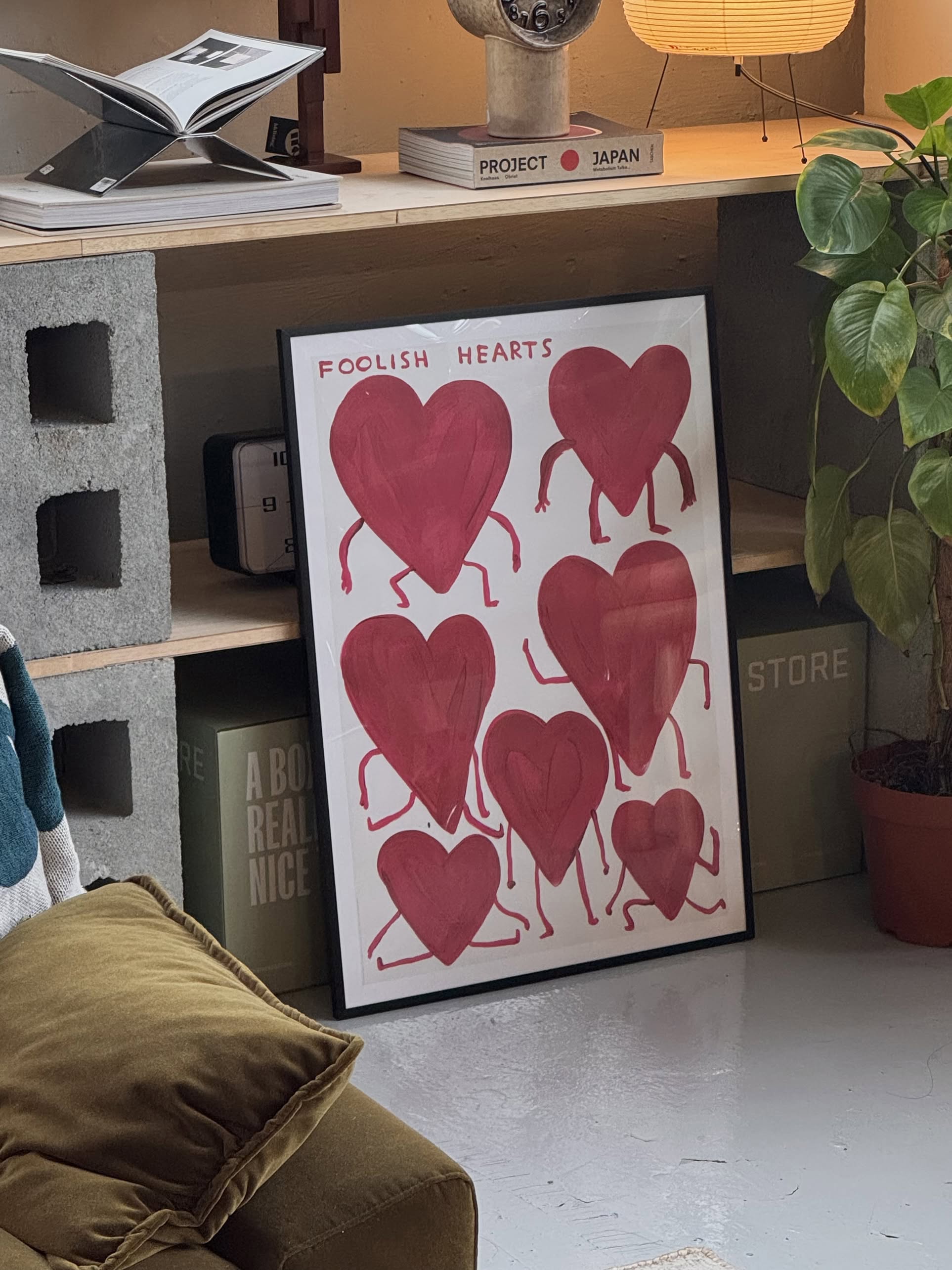 From Shrig Shop, the Foolish Hearts (50x70) framed artwork inspired by David Shrigley shows eight red, heart-shaped figures with arms and legs in a cozy modern room with books and plants.