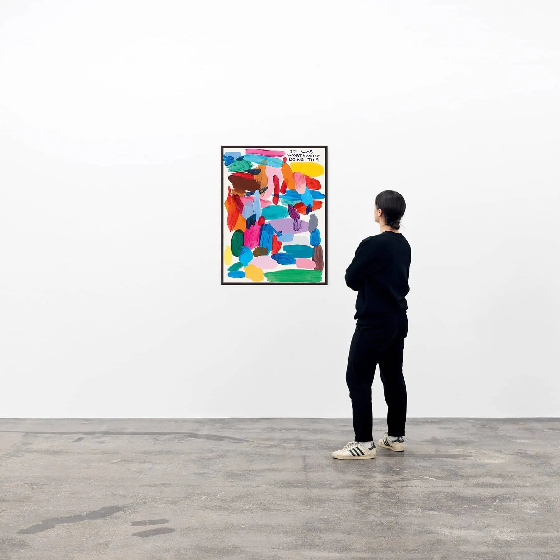 A person in black stands in a gallery, viewing It Was Worthwhile Doing This (60x80) by Shrig Shop—a David Shrigley poster featuring bold, overlapping red, blue, pink, green, and yellow shapes on a white wall.