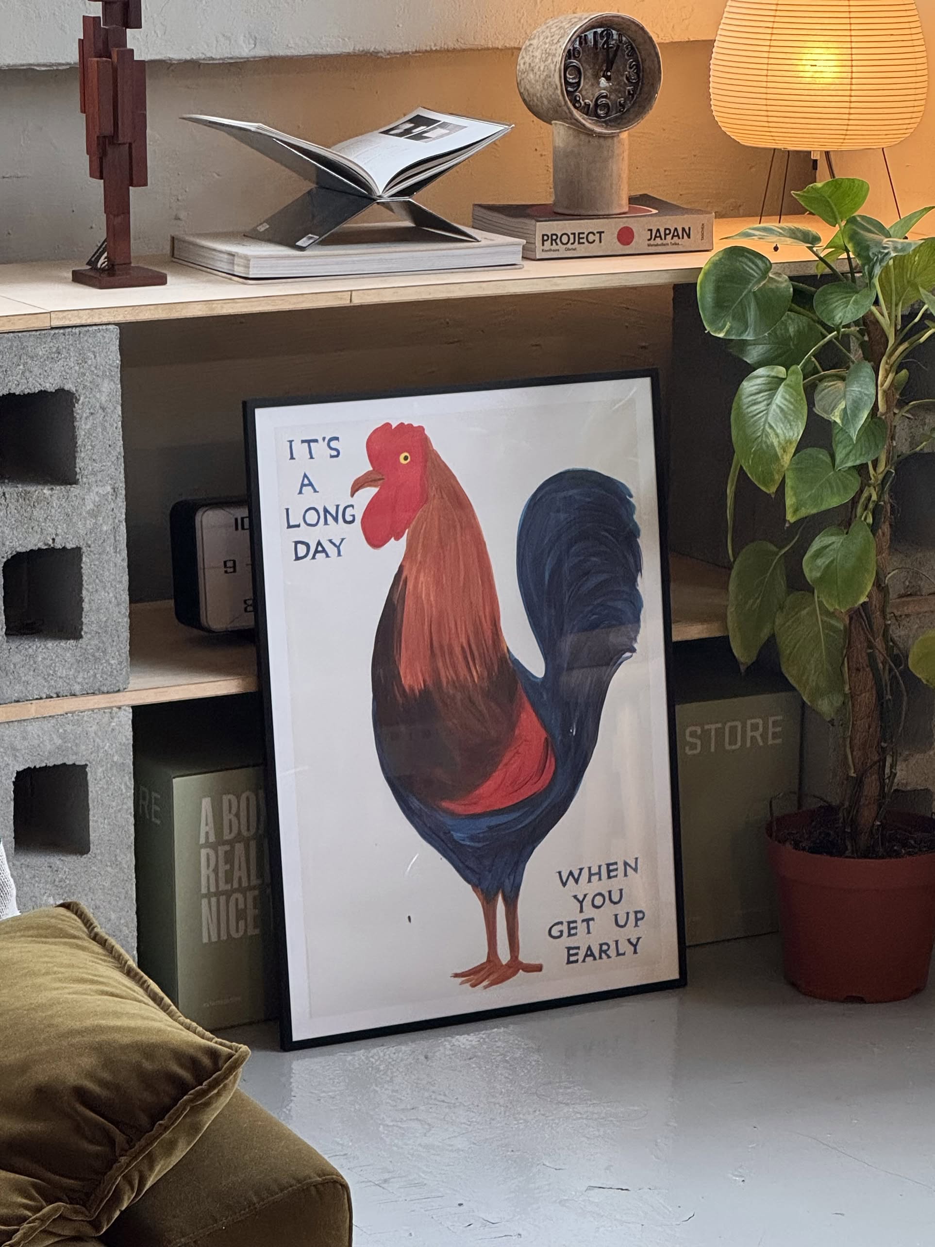 The Shrig Shop David Shrigley - Its A Long Day (50x70) framed print, featuring a rooster and the witty text “It’s a long day when you get up early,” leans against a shelf with books, plants, a clock, and lamp in a cozy room.
