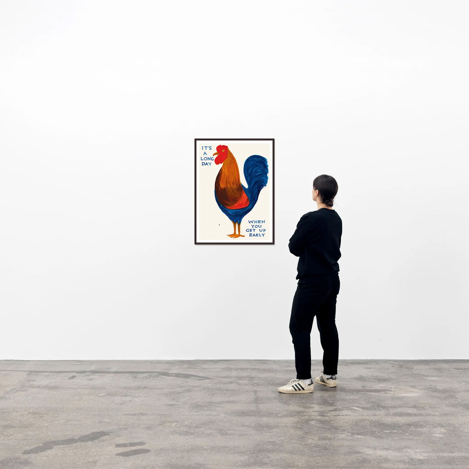 A person in black clothes and white sneakers views the Shrig Shop David Shrigley - Its A Long Day (50x70) poster, featuring a rooster and text, displayed on a white gallery wall.
