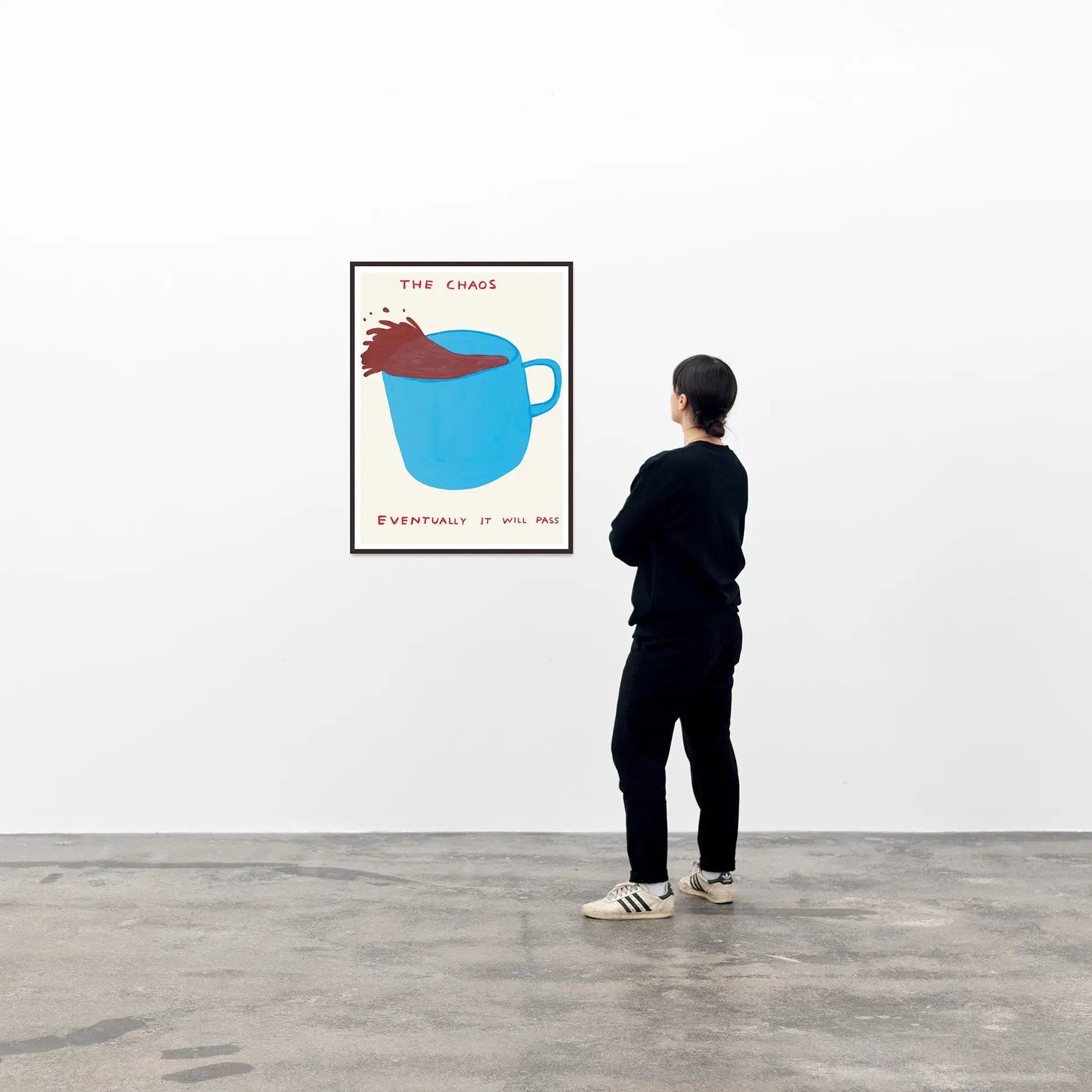 A person in black stands in a gallery, looking at Shrig Shop’s David Shrigley - The Chaos (60x80)—a poster of a blue mug with liquid spilling and the words THE CHAOS and EVENTUALLY IT WILL PASS.