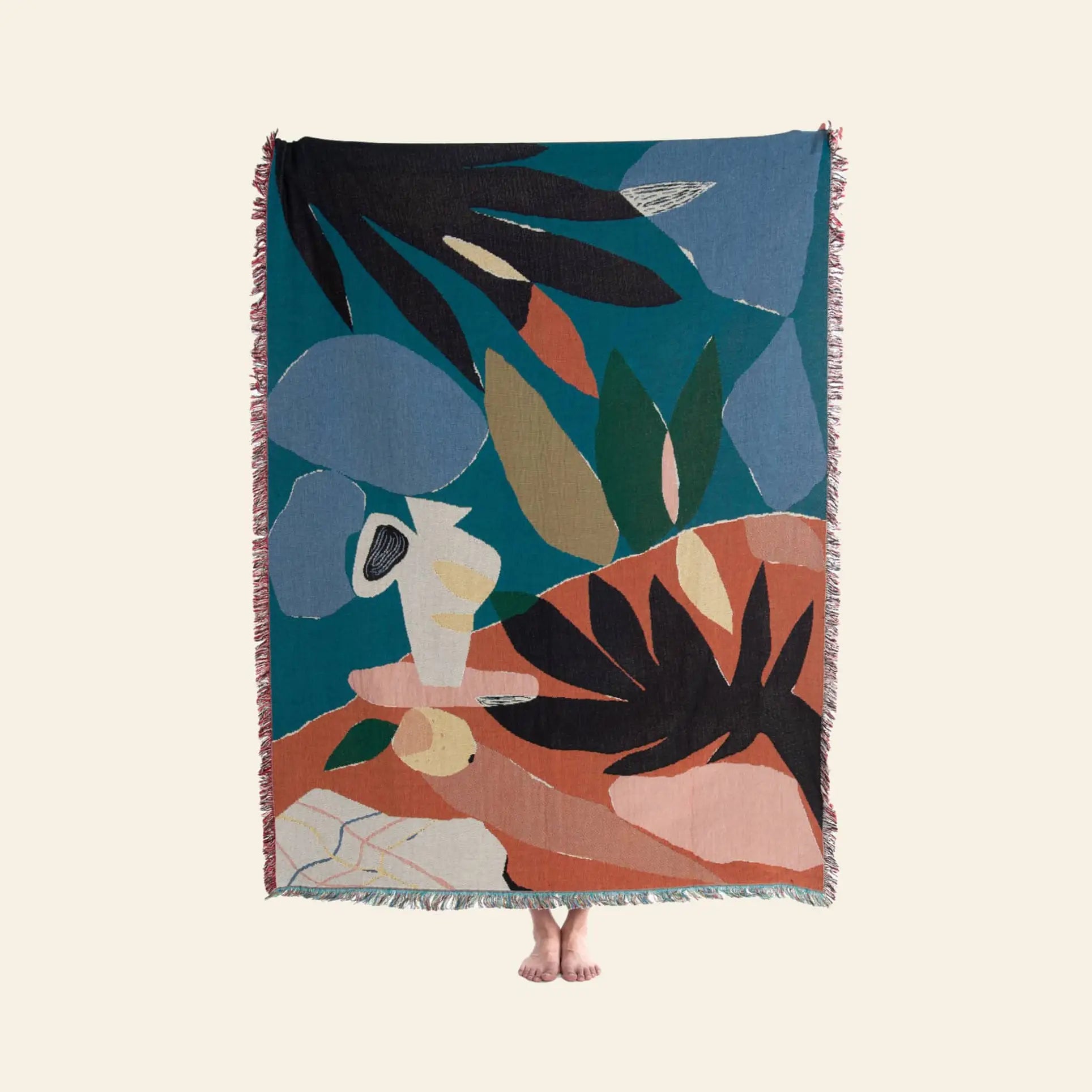 A person holds up the Slowdown Studio Barbosa Throw, featuring abstract plant-like patterns in blue, orange, green, and beige. Woven from recycled cotton by Slowdown Studio, only their bare feet are visible below the colorful blanket.