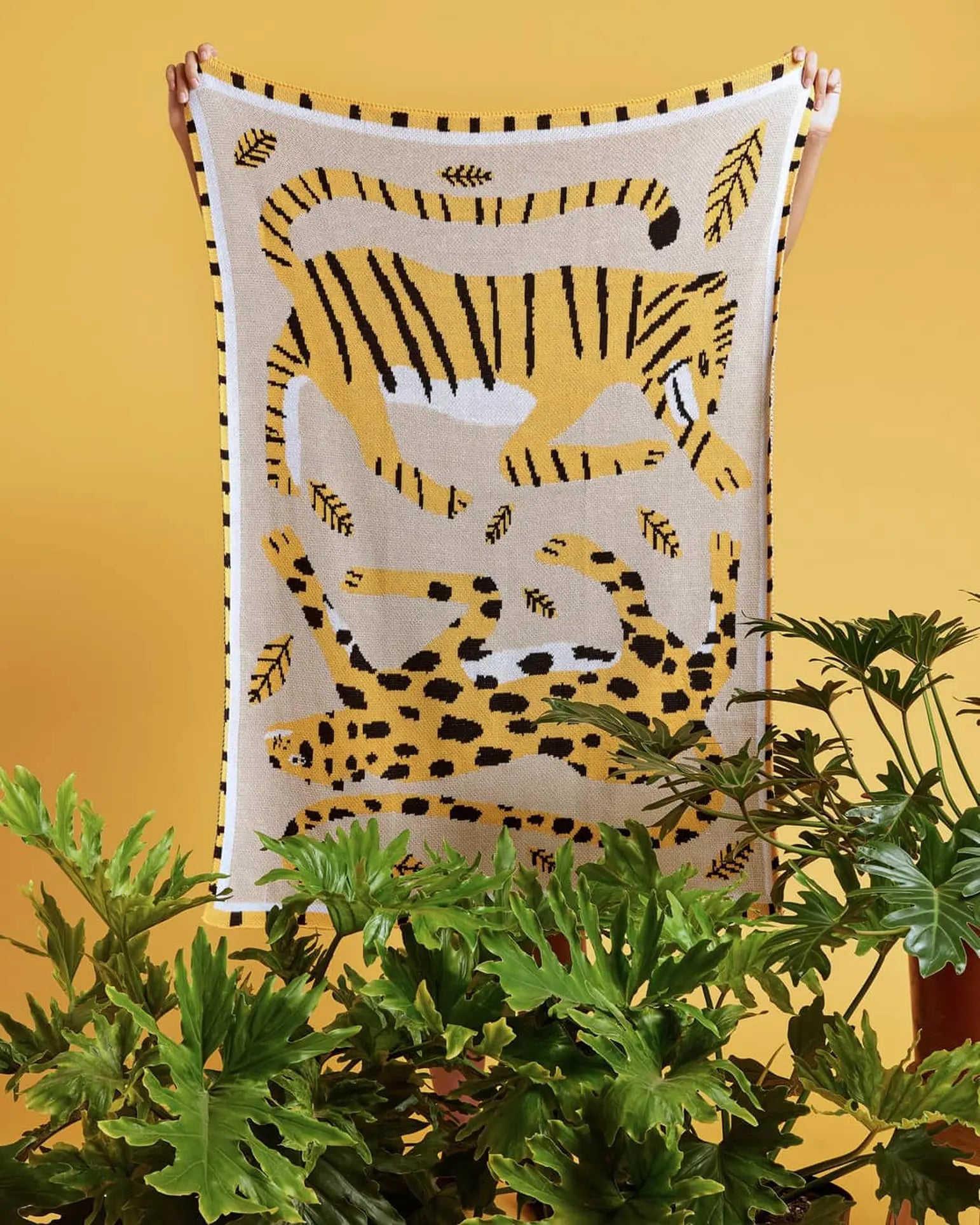 A person holds up the Slowdown Studio Big Cats Mini Blanket, made from a recycled cotton blend and showcasing abstract yellow, black, and white tiger and leopard designs with leaves on a striking yellow background.