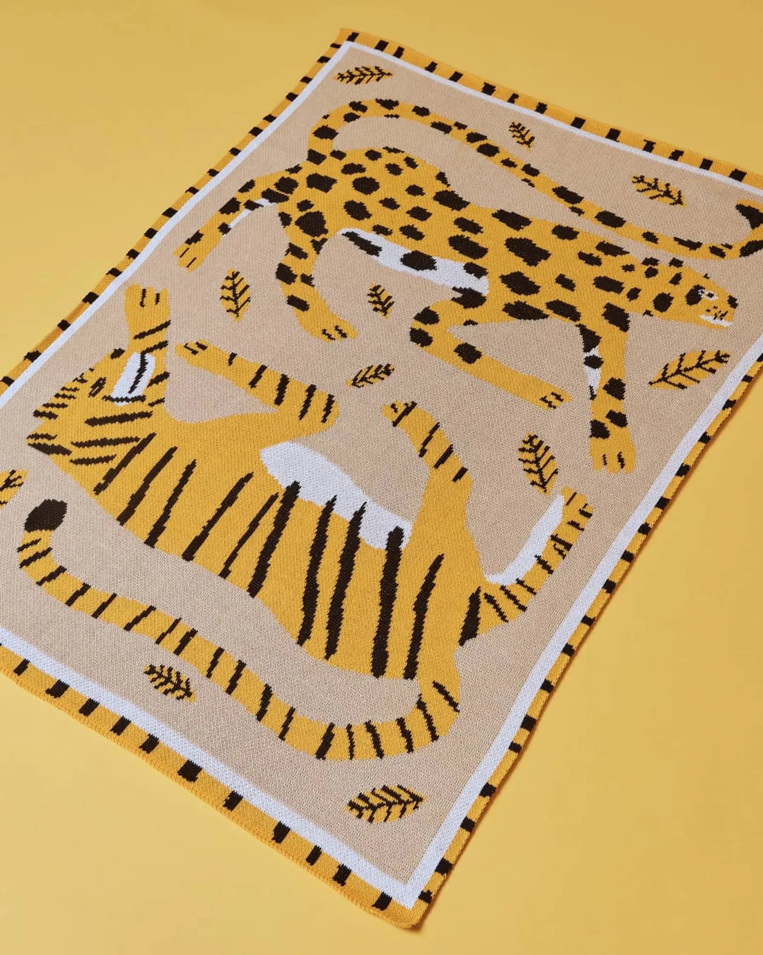 The Slowdown Studio Big Cats Mini Blanket is a rectangular beige recycled cotton blend with illustrated tigers, cheetahs, black and yellow leaves, a striped border, and a lively design on a yellow background.