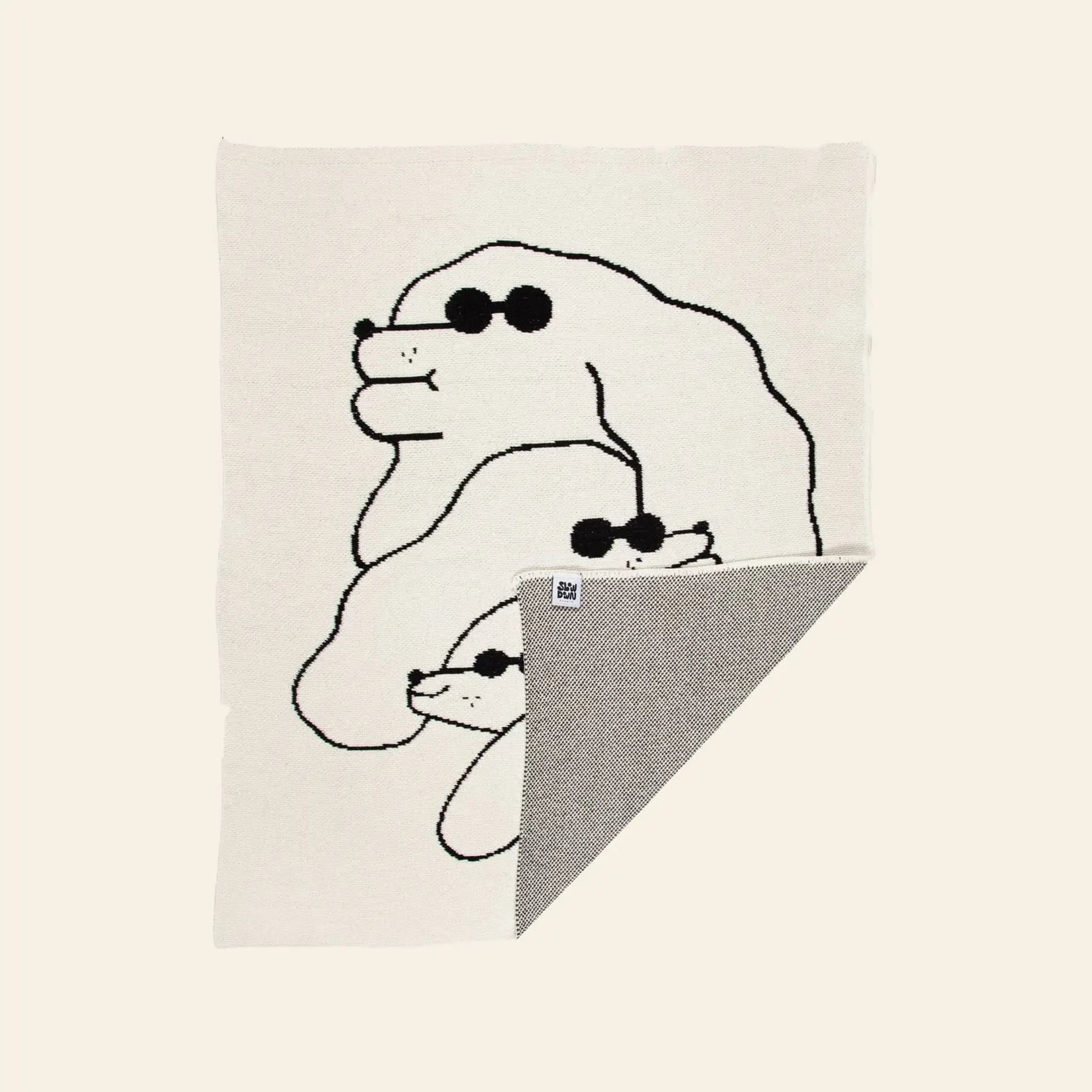 The Slowdown Studio Dawgs Mini Blanket is a cream-colored recycled cotton throw featuring a black line drawing of three dogs in sunglasses, stacked by size, with a folded corner showing the contrasting gray underside.