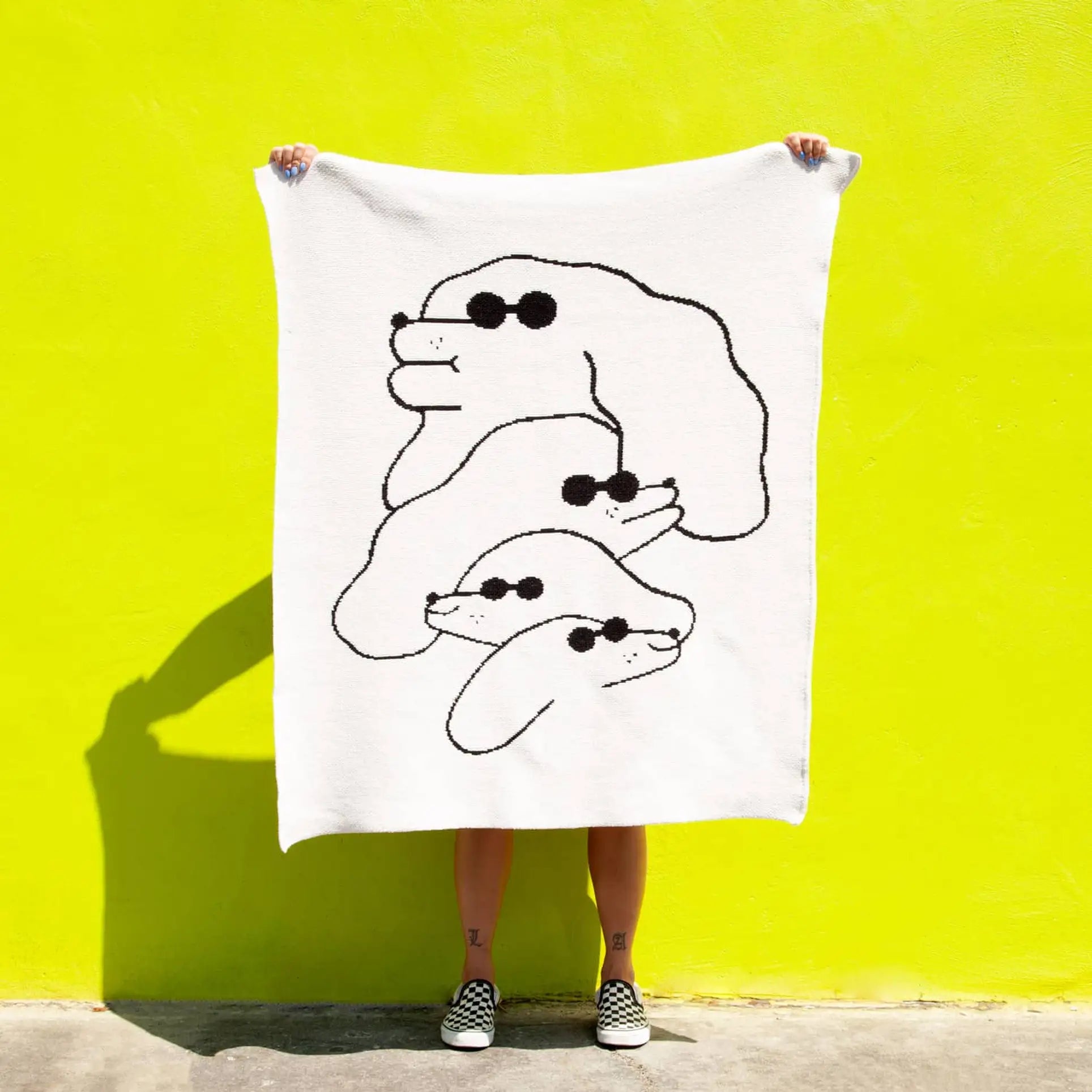A person stands against a bright yellow wall, holding up the Slowdown Studio Dawgs Mini Blanket—crafted from recycled cotton and featuring a minimalist black line drawing of three cartoon dogs in sunglasses. Only their legs and checkered shoes are visible.