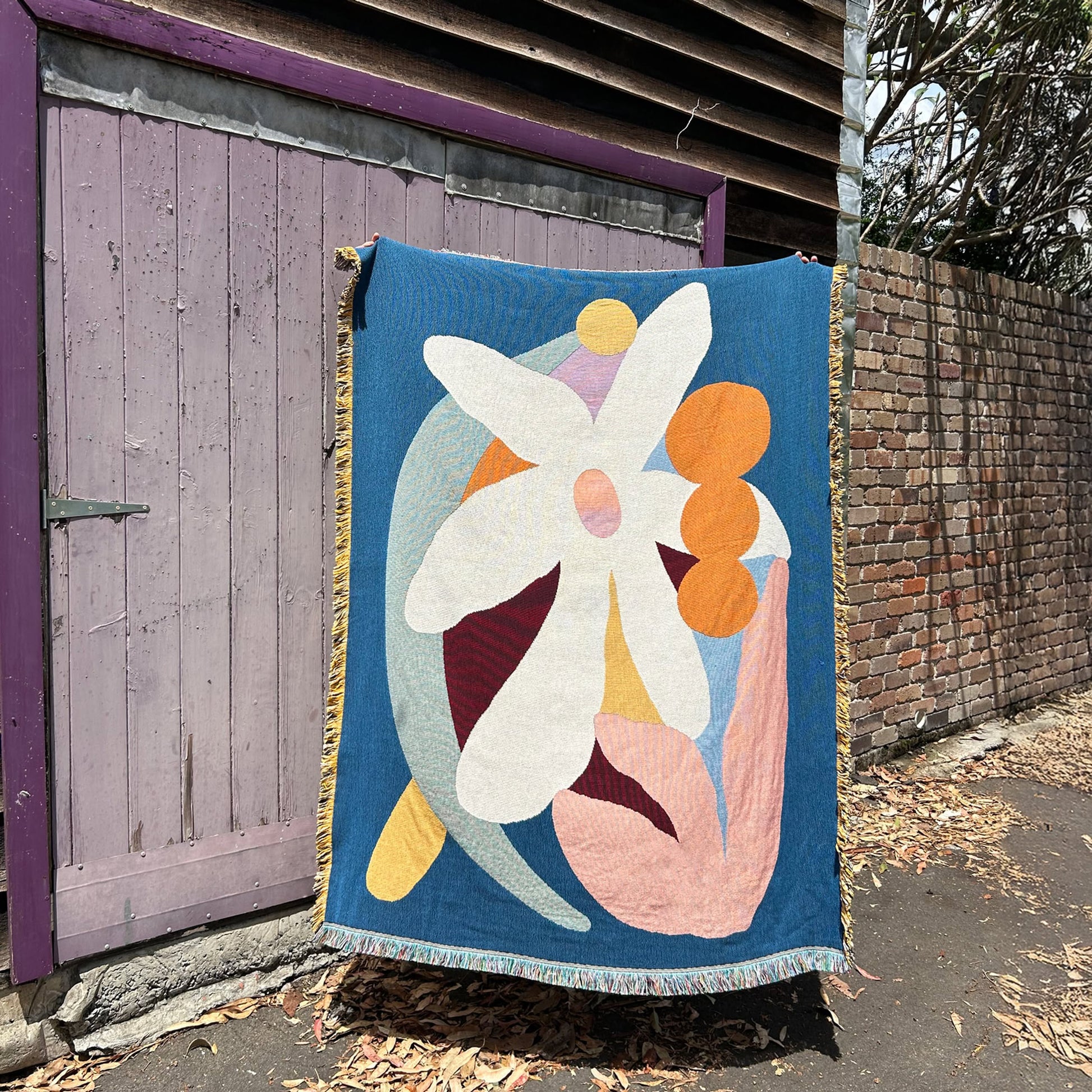 The Slowdown Studio Delaney Throw, made from recycled cotton with an abstract floral design, hangs outdoors before a weathered purple wooden door and brick wall, dry leaves scattered on the ground.