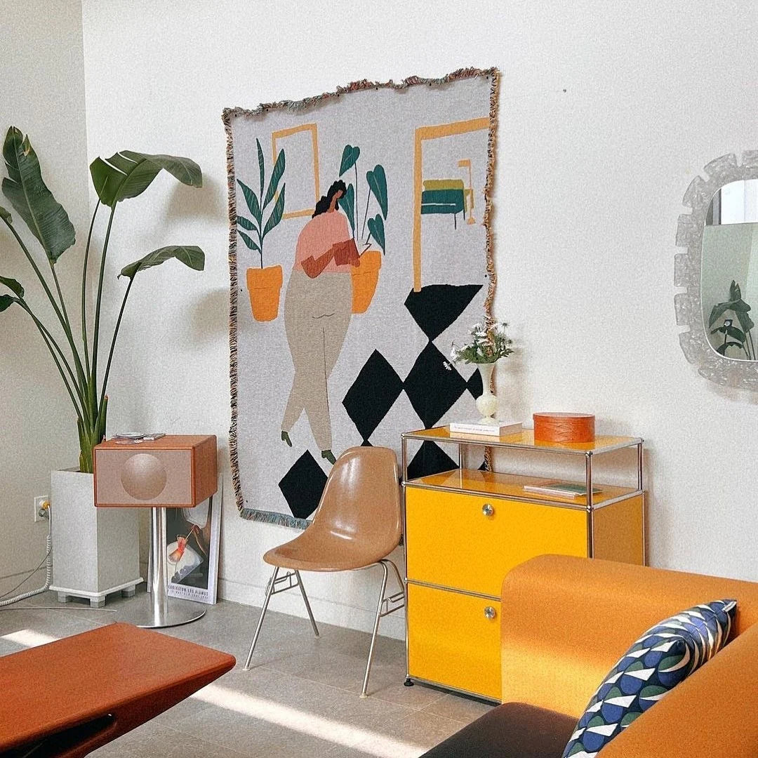 A colorful, modern living room features a large plant, brown chair, orange and yellow cabinets, a small side table, decorative mirror, and the Slowdown Studio Dresden Throw by Slowdown Studio—a versatile abstract art tapestry made from recycled cotton. Sunlight brightens the cozy space.