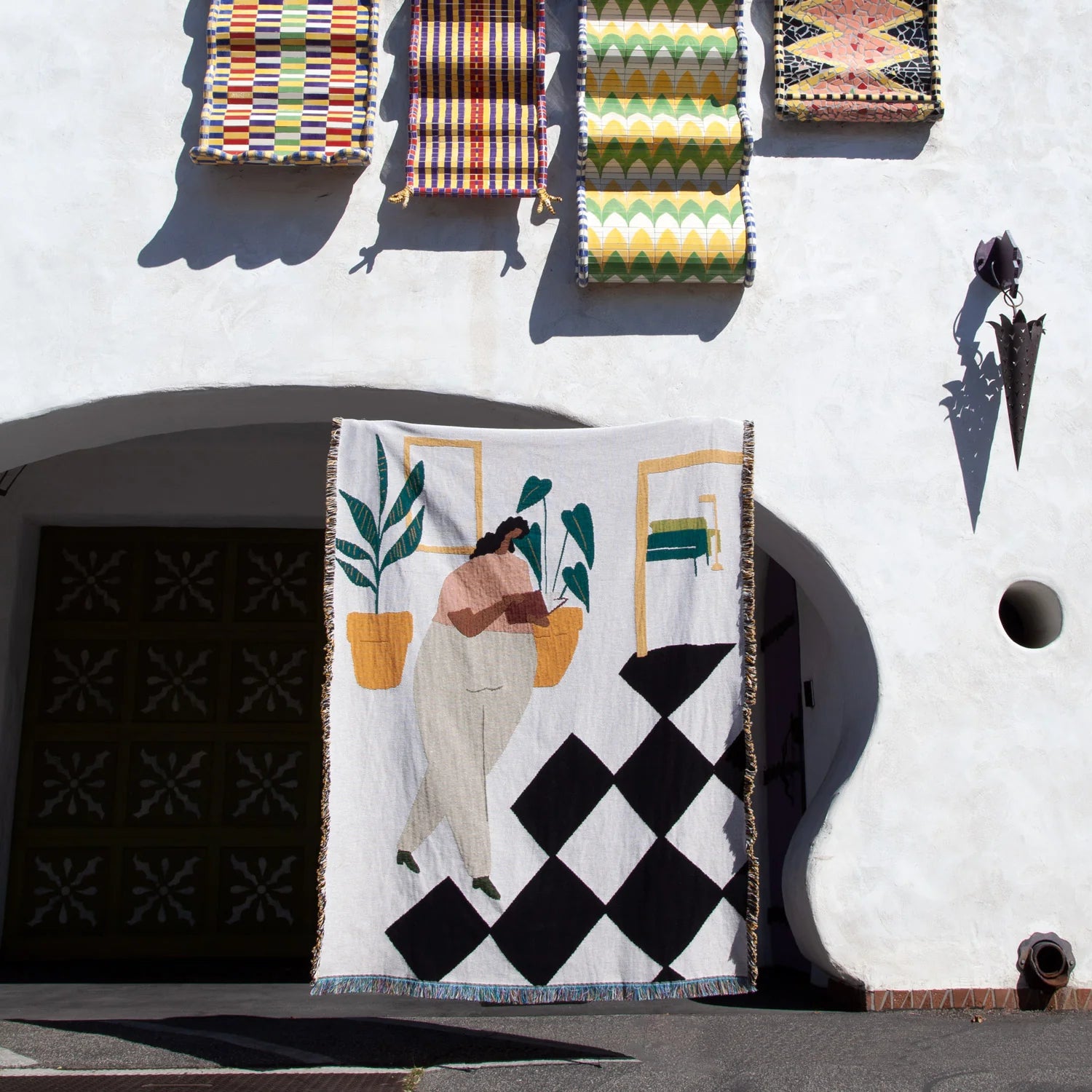The Slowdown Studio Dresden Throw by Slowdown Studio, a recycled cotton blanket with an abstract figure, plants, and geometric designs, hangs outside a white building beneath four vibrant rugs displayed above a dark arched doorway.