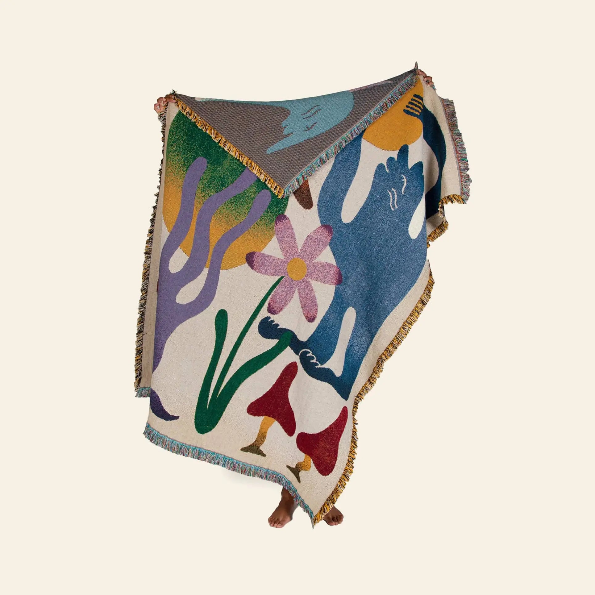 Wrapped in the Slowdown Studio Dumont Throw, a cozy woven blanket featuring abstract shapes, a blue figure, hands, a large flower, and red mushrooms on cream recycled cotton, only the persons feet peek out below.