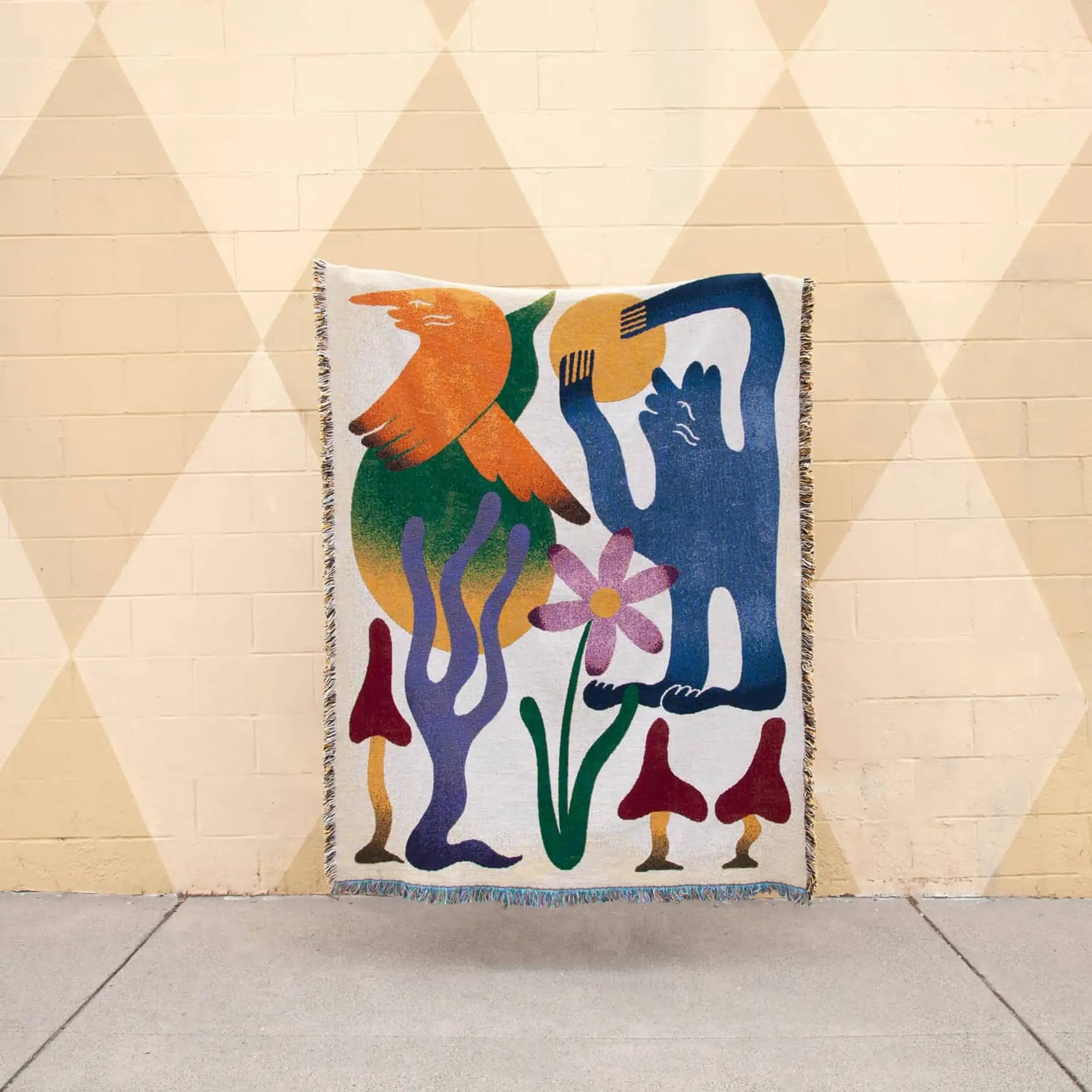The Slowdown Studio Dumont Throw is a woven blanket made from recycled cotton, featuring abstract, colorful shapes including a blue figure, orange bird, pink flower, red mushrooms, and purple coral on a beige diamond-patterned background.