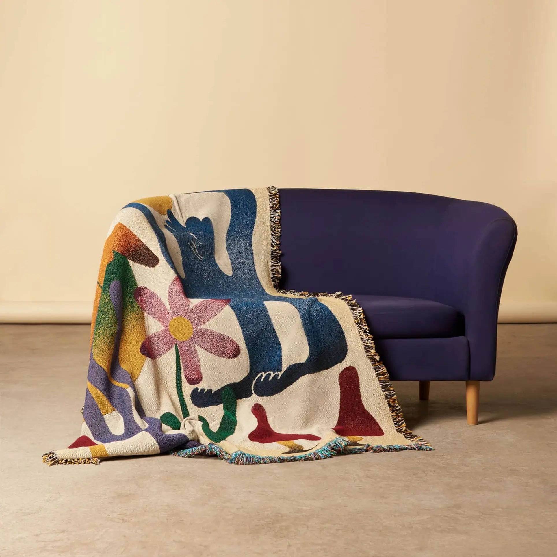 The Slowdown Studio Dumont Throw, a colorful recycled cotton blanket with abstract shapes and a bold pink flower, is draped over a modern dark blue loveseat on a light floor against a beige wall.