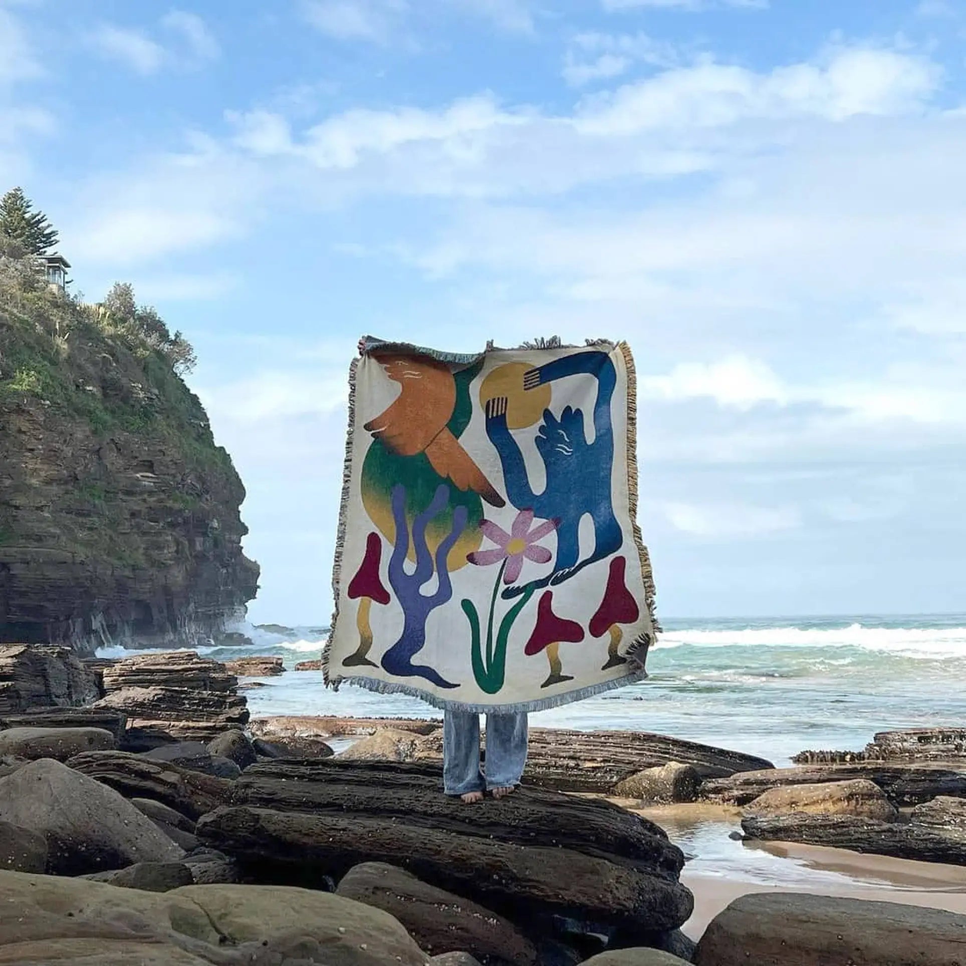 A person stands on a rocky shore, holding up the Slowdown Studio Dumont Throw—a colorful blanket by Slowdown Studio featuring nature-inspired shapes—while a cliff and the ocean are visible beneath a partly cloudy sky.