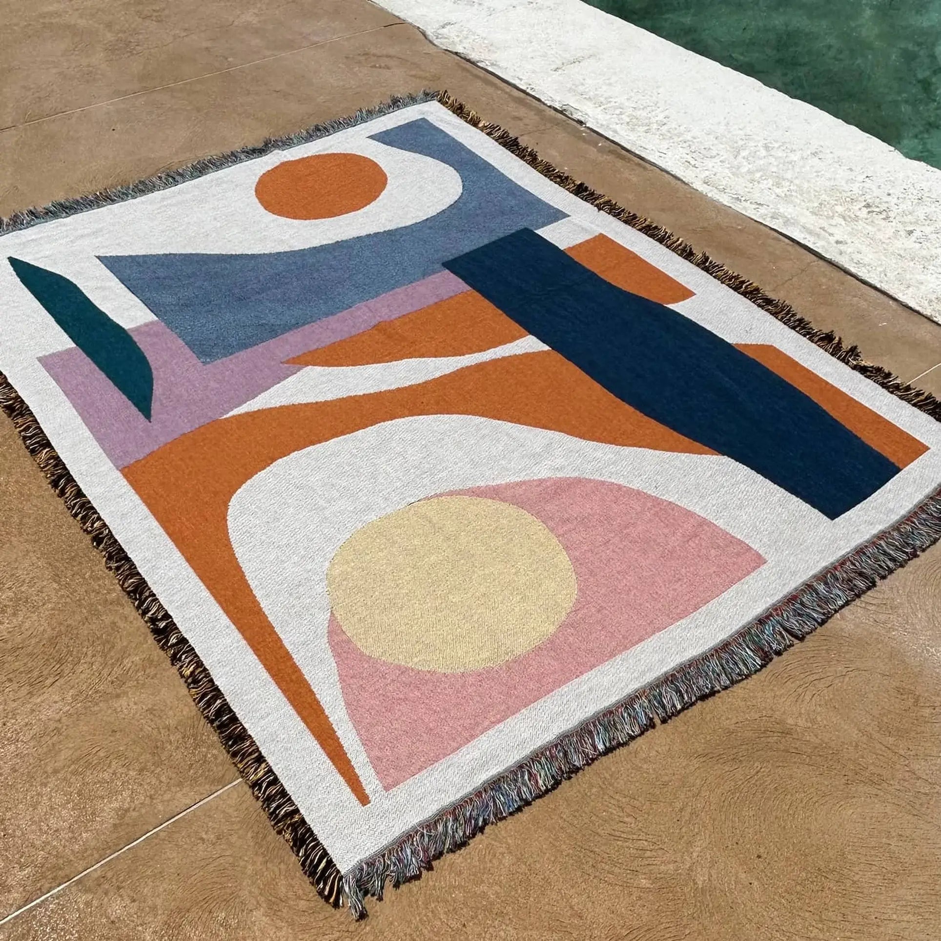 The Slowdown Studio Essien Throw, made from recycled cotton and featuring abstract blue, orange, pink, and cream shapes with a fringe border, rests on a textured brown surface by a pool with clear green water.