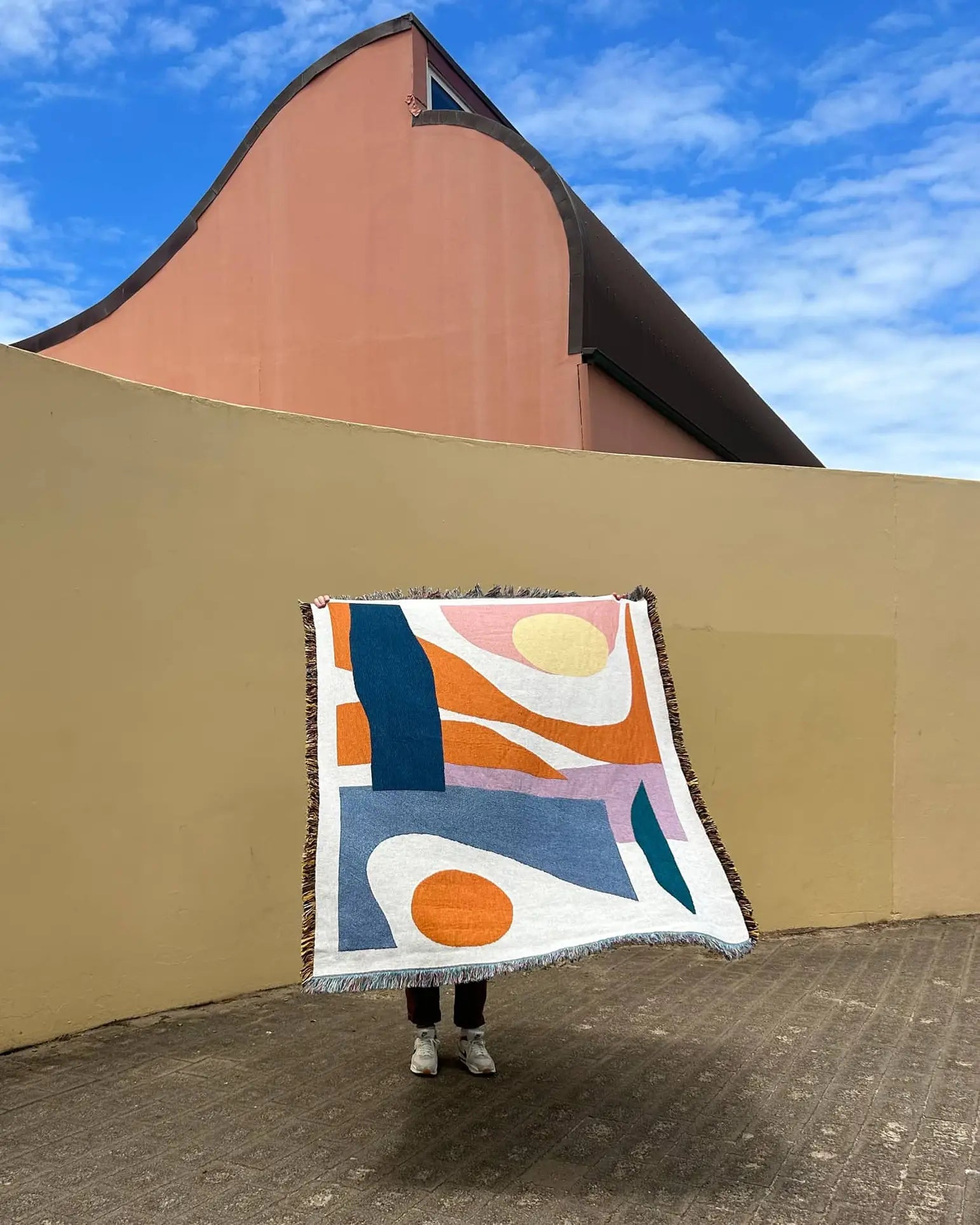 A person stands holding the Slowdown Studio Essien Throw—a colorful, abstract-patterned woven blanket—obscuring their body in front of a peach building and beige wall beneath a blue sky with scattered clouds.
