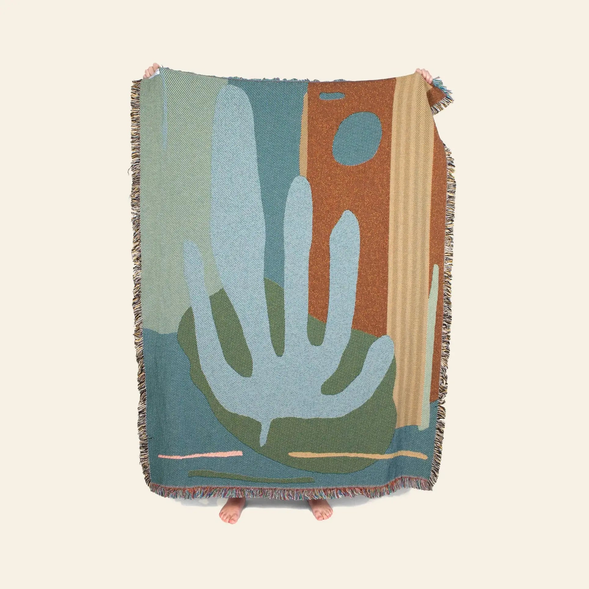A person holds up the Slowdown Studio Hazlewood Throw, an eco-friendly woven blanket by Slowdown Studio with abstract blue, green, and rust shapes. Only their hands and feet appear behind it against a plain background.