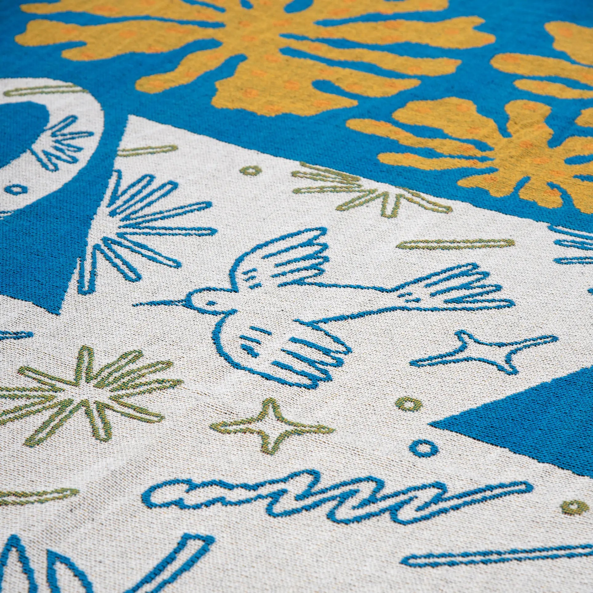 A close-up of the Slowdown Studio Hudson Throw shows colorful embroidery—blue bird, green and yellow stars, abstract designs, and orange florals—on blue and white recycled cotton by Slowdown Studio.
