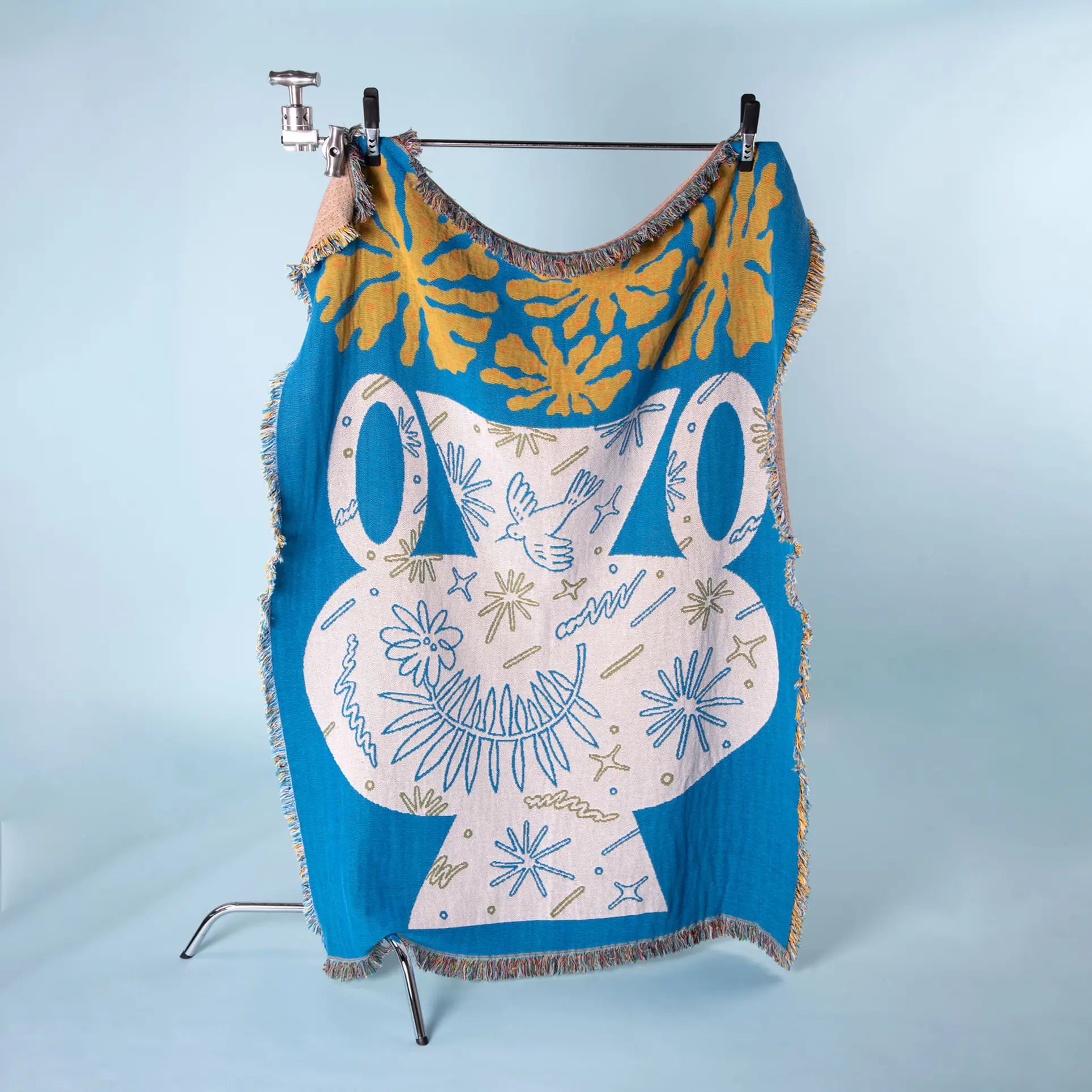 The Slowdown Studio Hudson Throw, featuring an abstract frog and patterns in blue and yellow recycled cotton, hangs on a metal rack against a light blue background, showcasing Slowdown Studios sustainable touch.