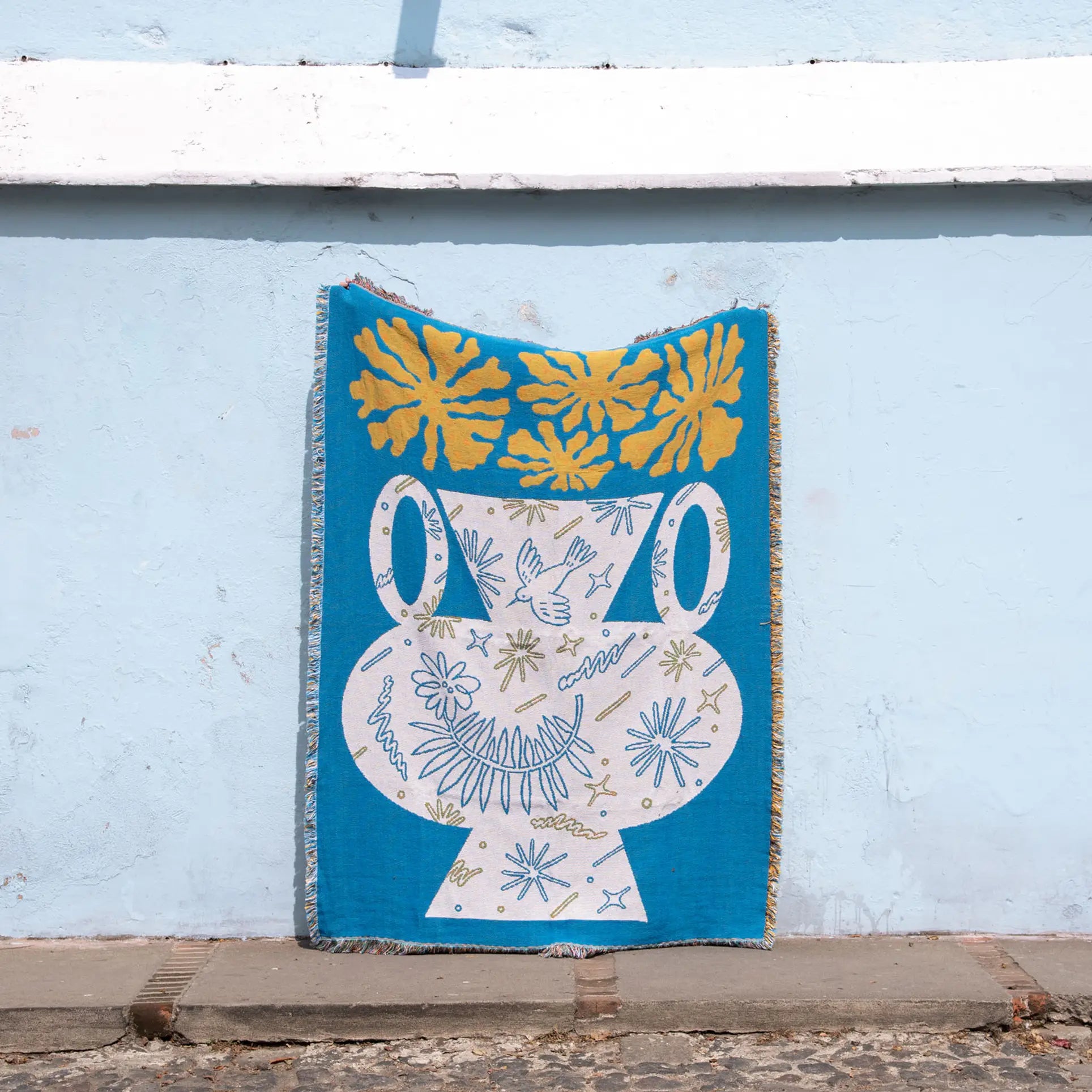 The Slowdown Studio Hudson Throw by Slowdown Studio is a blue blanket made from recycled cotton, depicting a stylized white vase with yellow leaves and abstract flowers. It hangs on a light blue wall above cobblestone flooring.