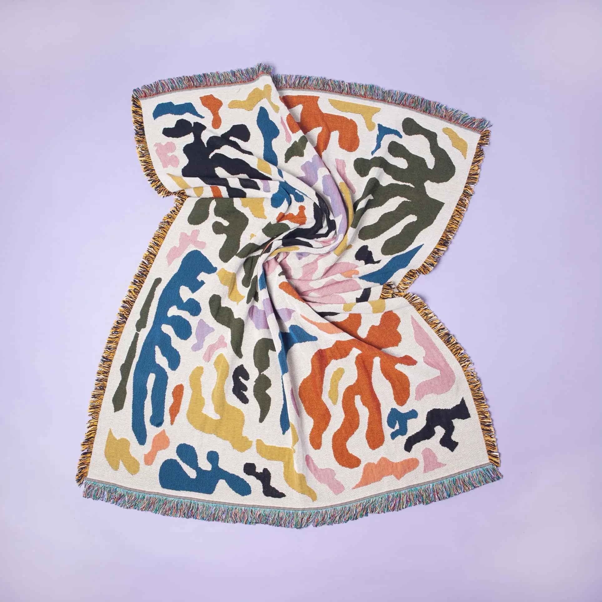 The Slowdown Studio Mercer Throw is a multicolored woven blanket made from recycled cotton, featuring abstract wavy patterns and fringed edges, twisted in the center and laid flat on a pale lavender surface.